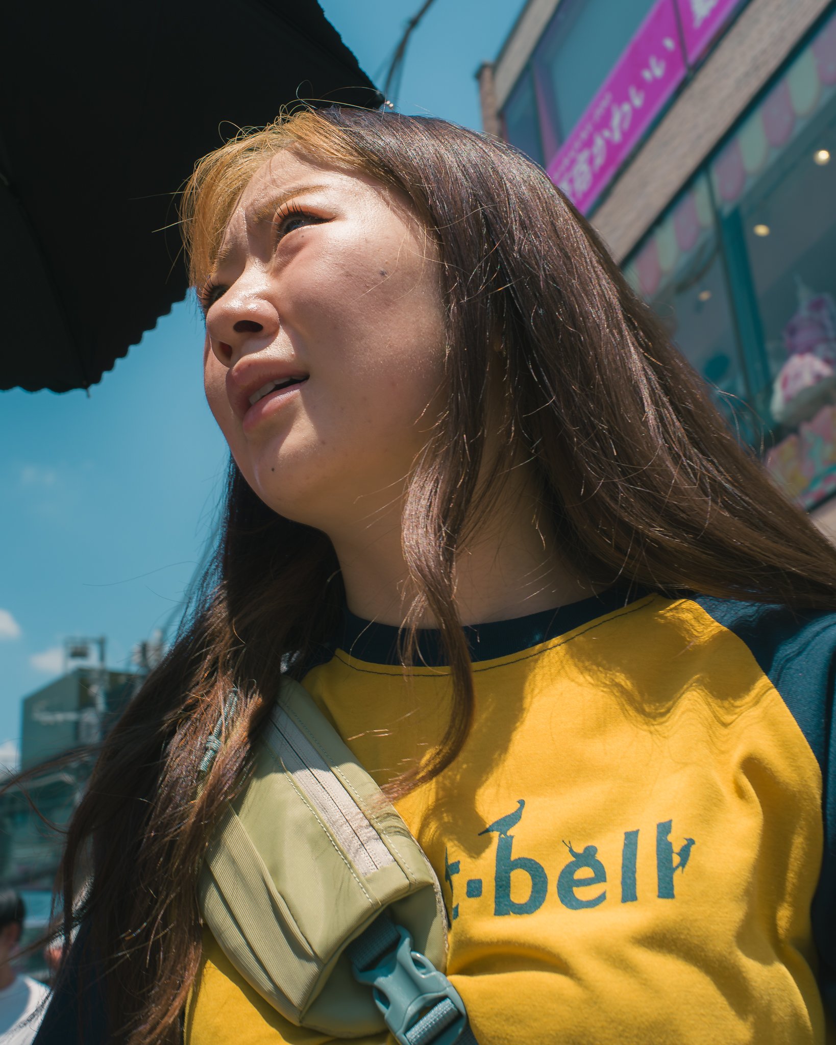 Close-up of a woman with long hair wearing a yellow shirt with blue text, with bright sunlight on her face, and a building with pink and blue signage in the background.