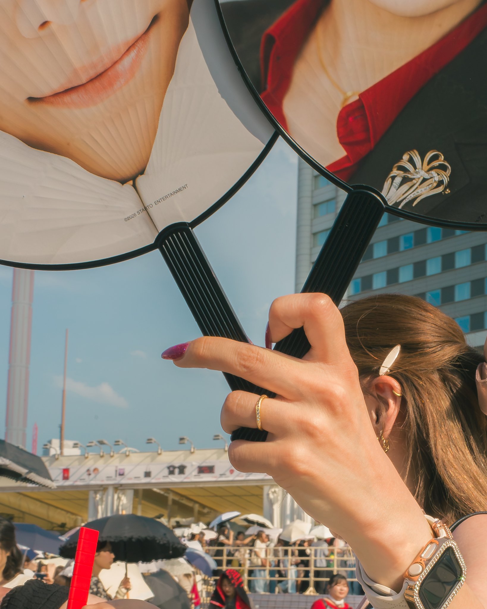 Close-up of a woman holding a mirror reflecting her face, skin, and makeup, with a crowd outdoors with umbrellas and buildings in the background.