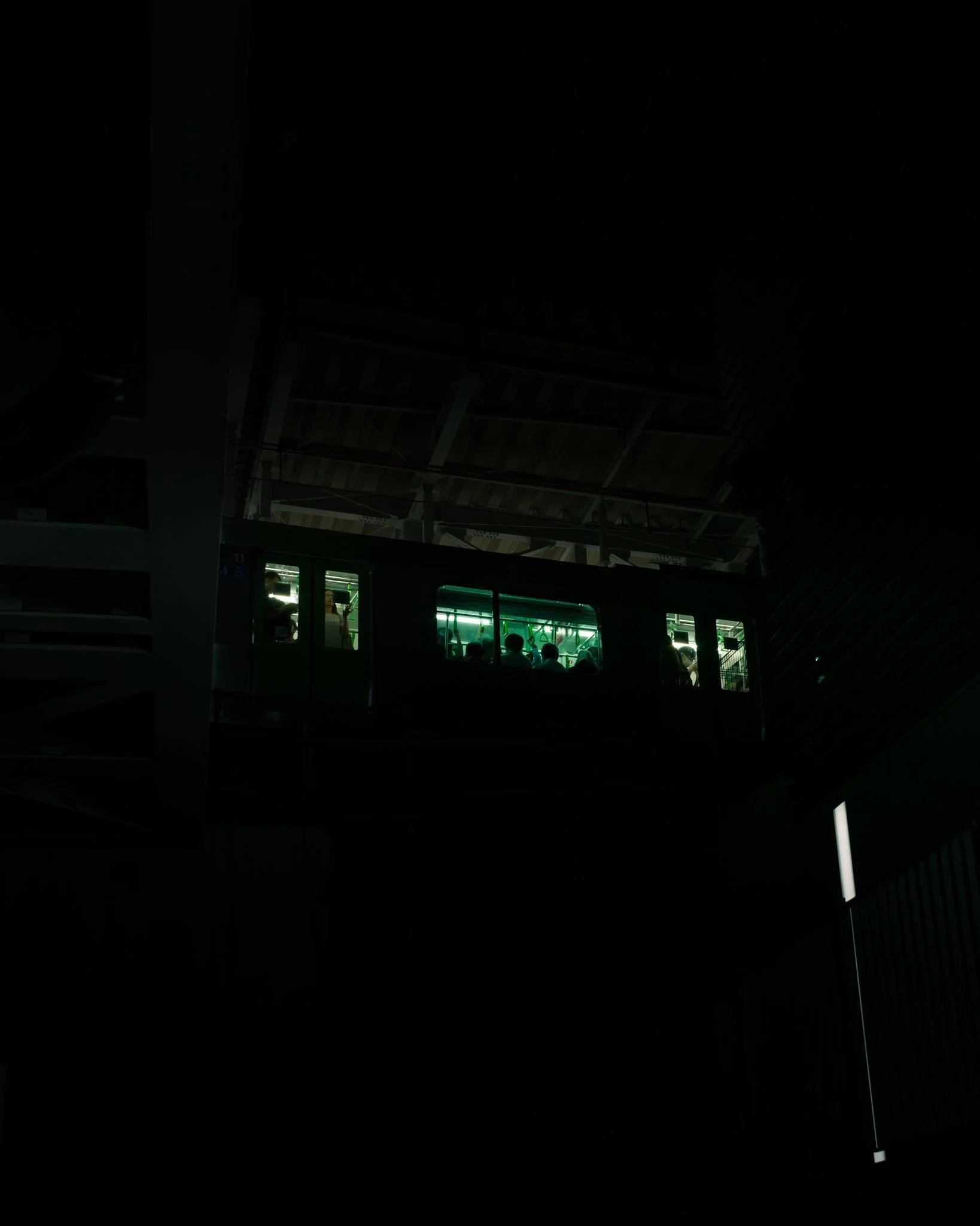 Dark interior scene of a building with a visible escalator and a glass elevator, passengers inside, with a bright green-lit storefront visible through the glass.
