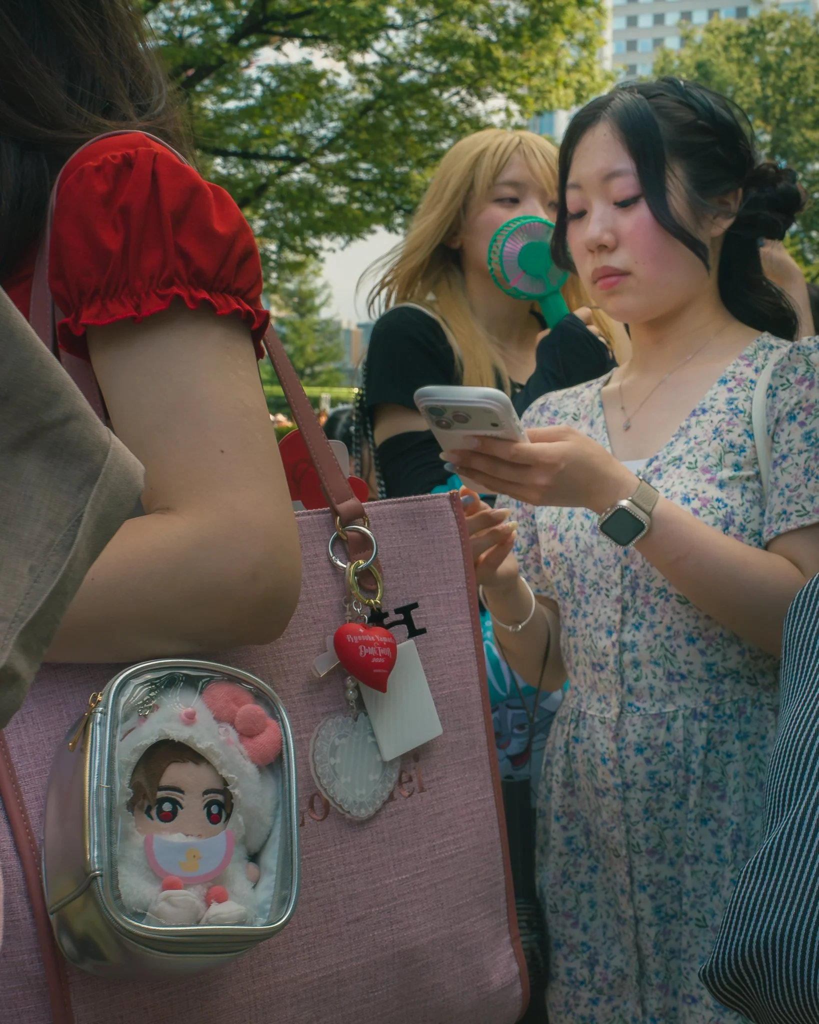 A group of young women gathered outdoors, one using a smartphone, with trees and a building in the background. One woman has a fan, and another has a small backpack with a plush doll attached.