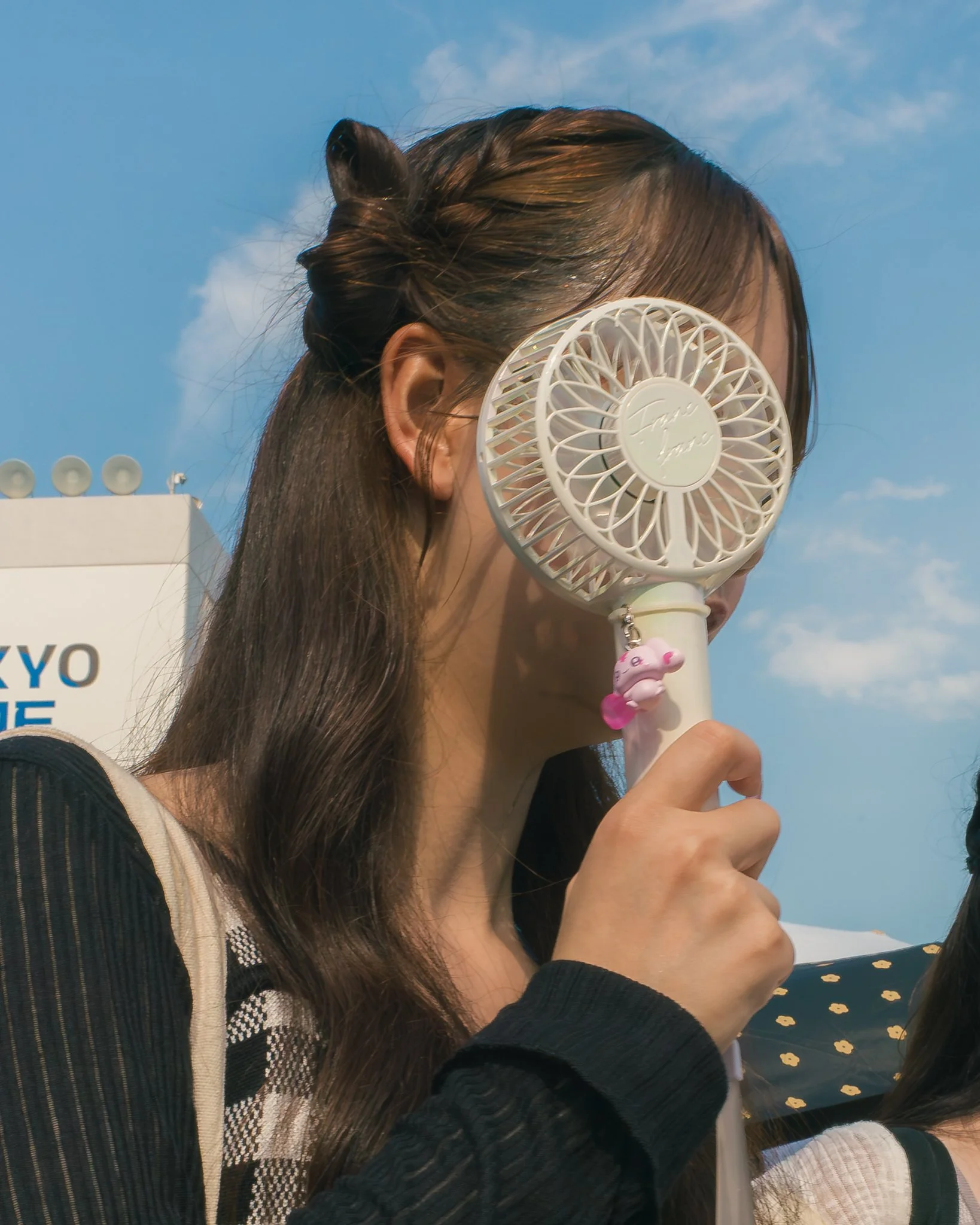 A woman with dark brown hair is holding a white handheld fan with a circular design near her face, outdoors on a sunny day with a blue sky and clouds.