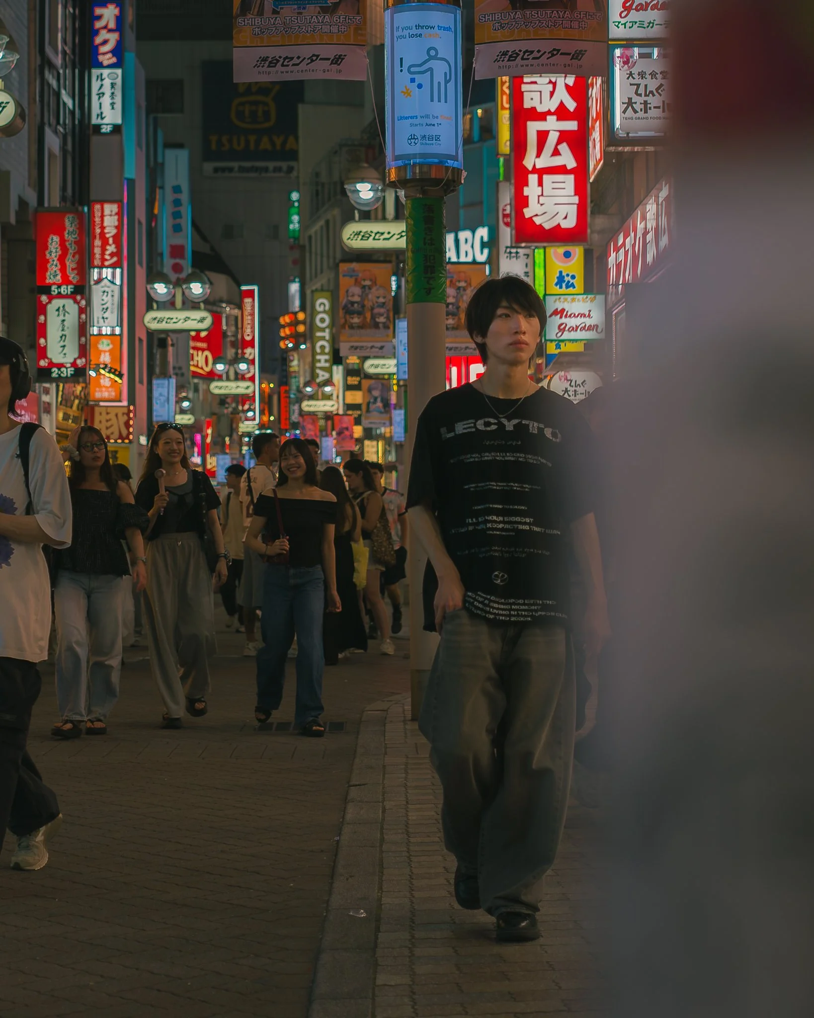 Nighttime street scene in Tokyo with neon signs in Japanese and English, pedestrians walking on the sidewalk, and a young man in the foreground wearing a black T-shirt and gray pants.