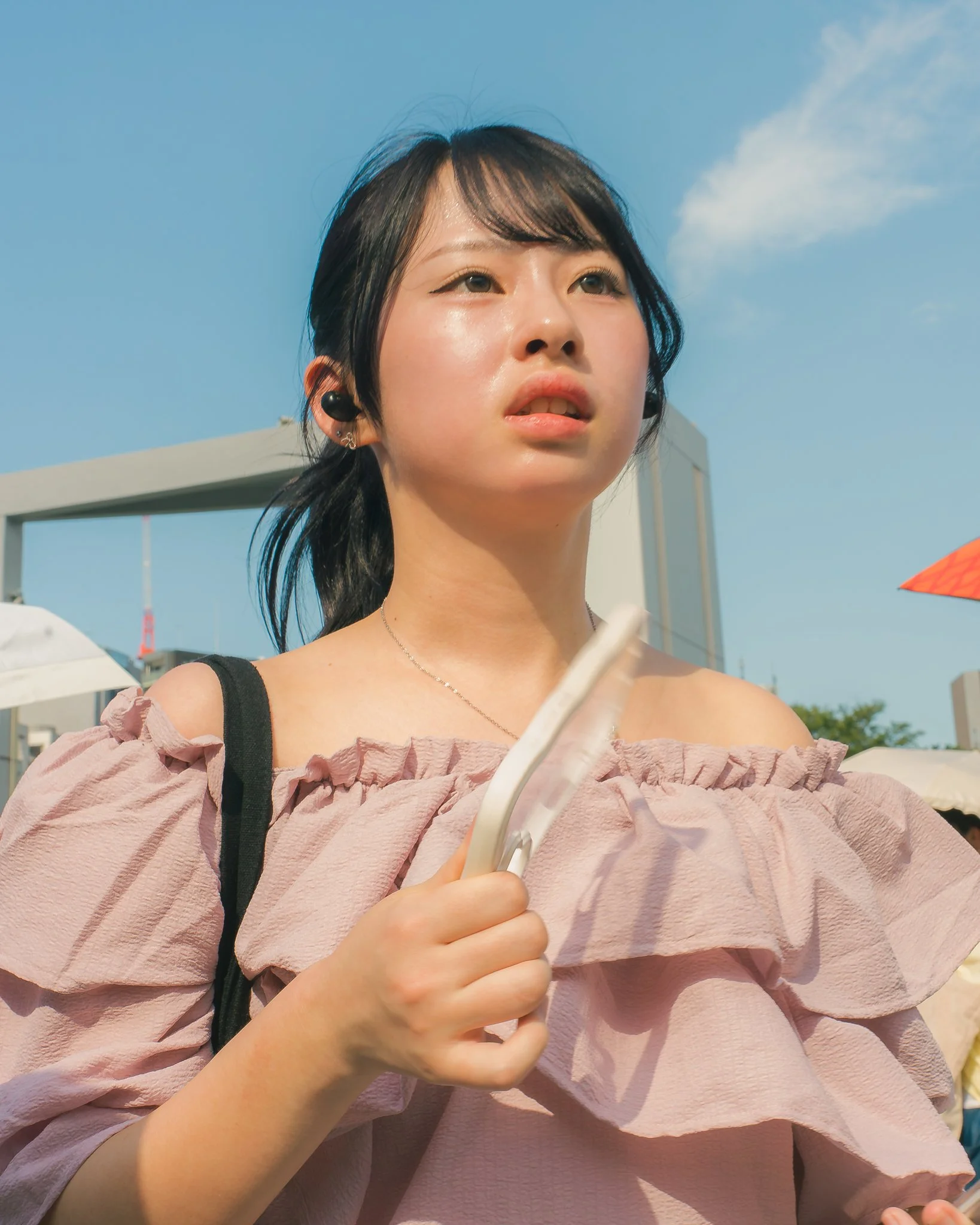 Close-up of a young woman with short black hair, wearing earrings and a pink ruffled top, holding a white smartphone outdoors against a clear blue sky.