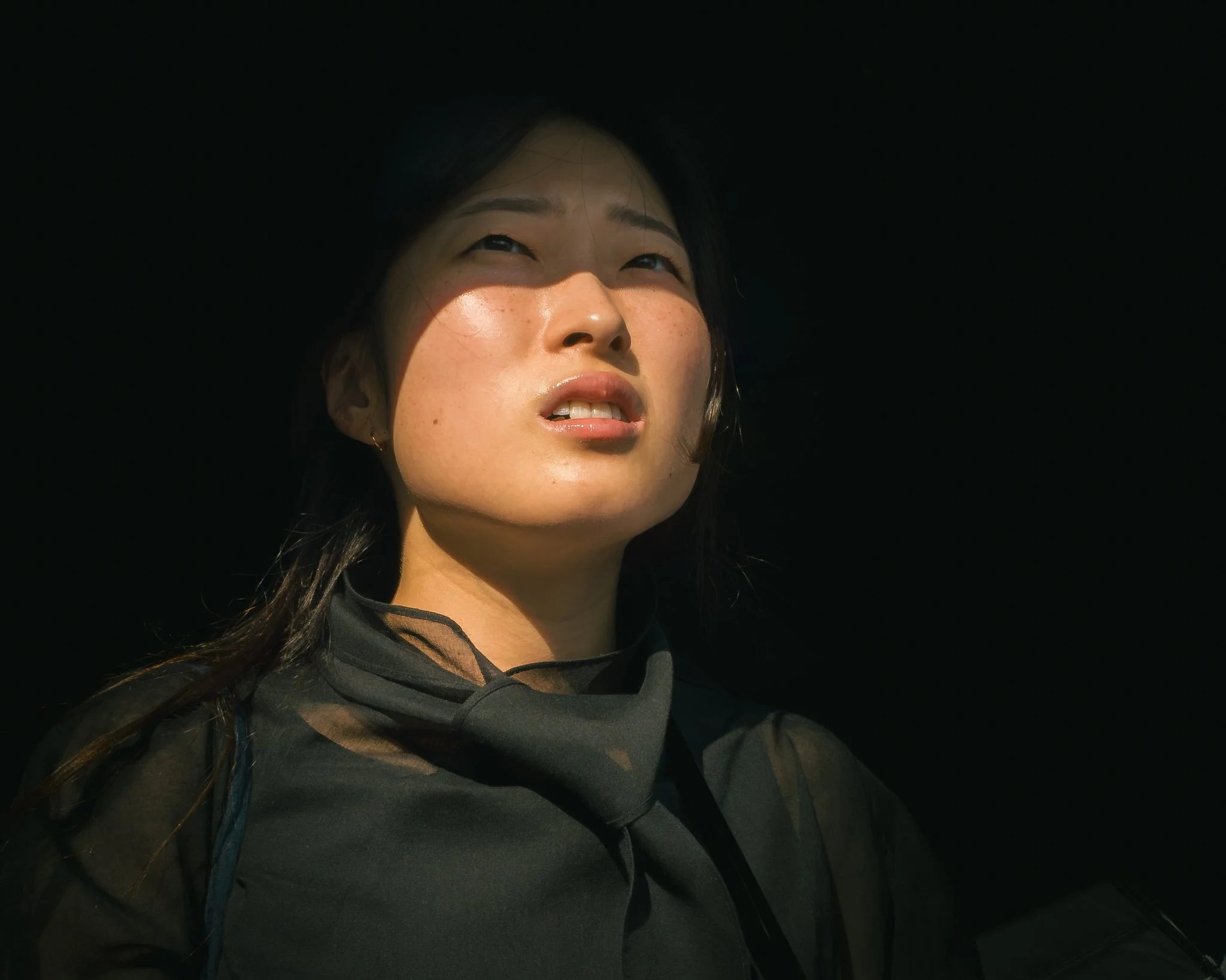 Close-up of a young woman looking upward with a subtle smile, dark long hair, and wearing a dark hoodie, with dramatic lighting highlighting her face against a dark background.