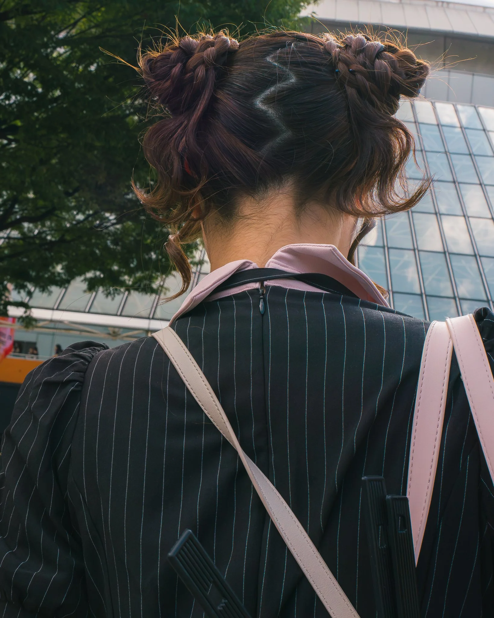 Back view of a woman with braided hairstyle, wearing a pinstripe jacket, carrying a pink bag, standing outdoors in front of a modern glass building and green trees.