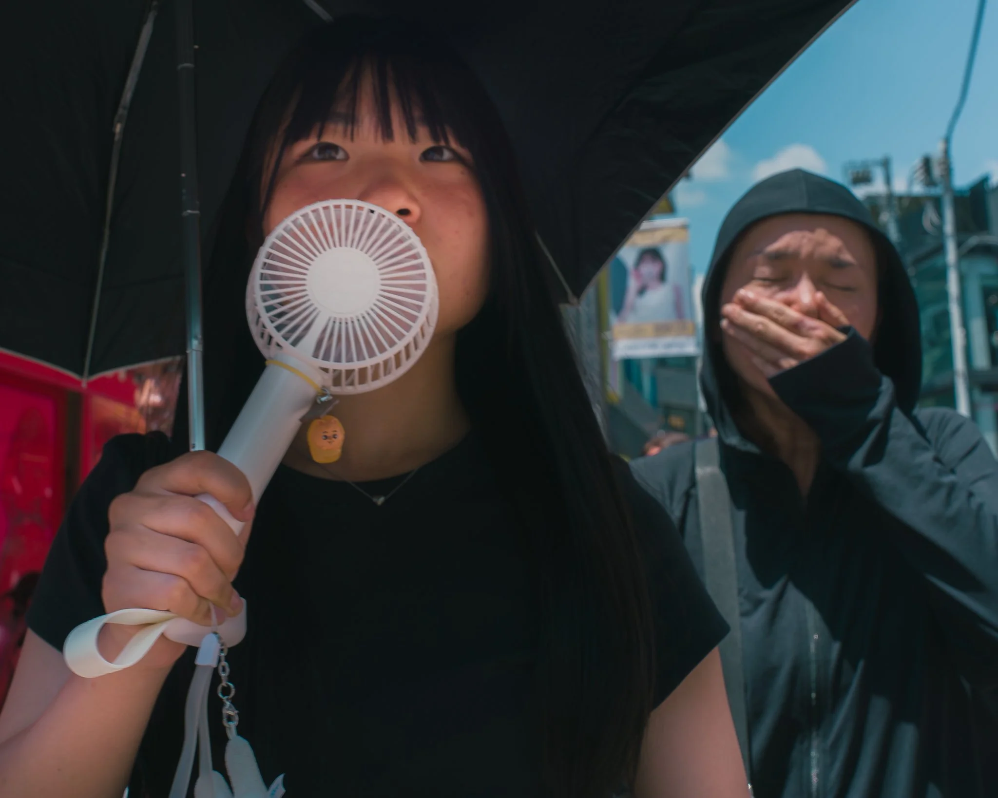 Close-up of a young woman holding a small portable fan near her face, with another woman in the background covering her mouth with her hand, both under an umbrella in an urban setting with bright blue sky.