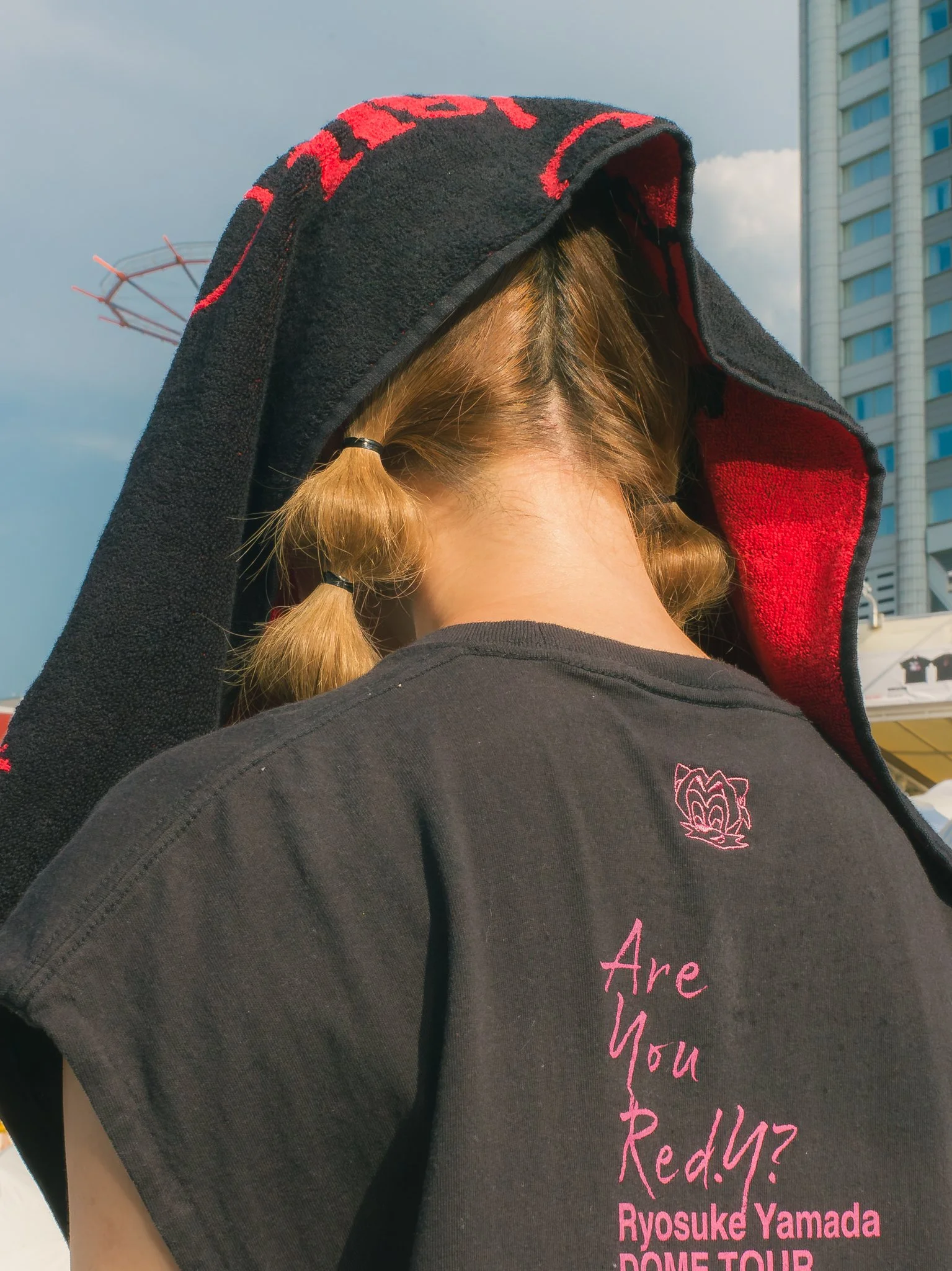 Close-up of a person wearing a black t-shirt with pink text, a towel overlaying their head, and a building in the background.