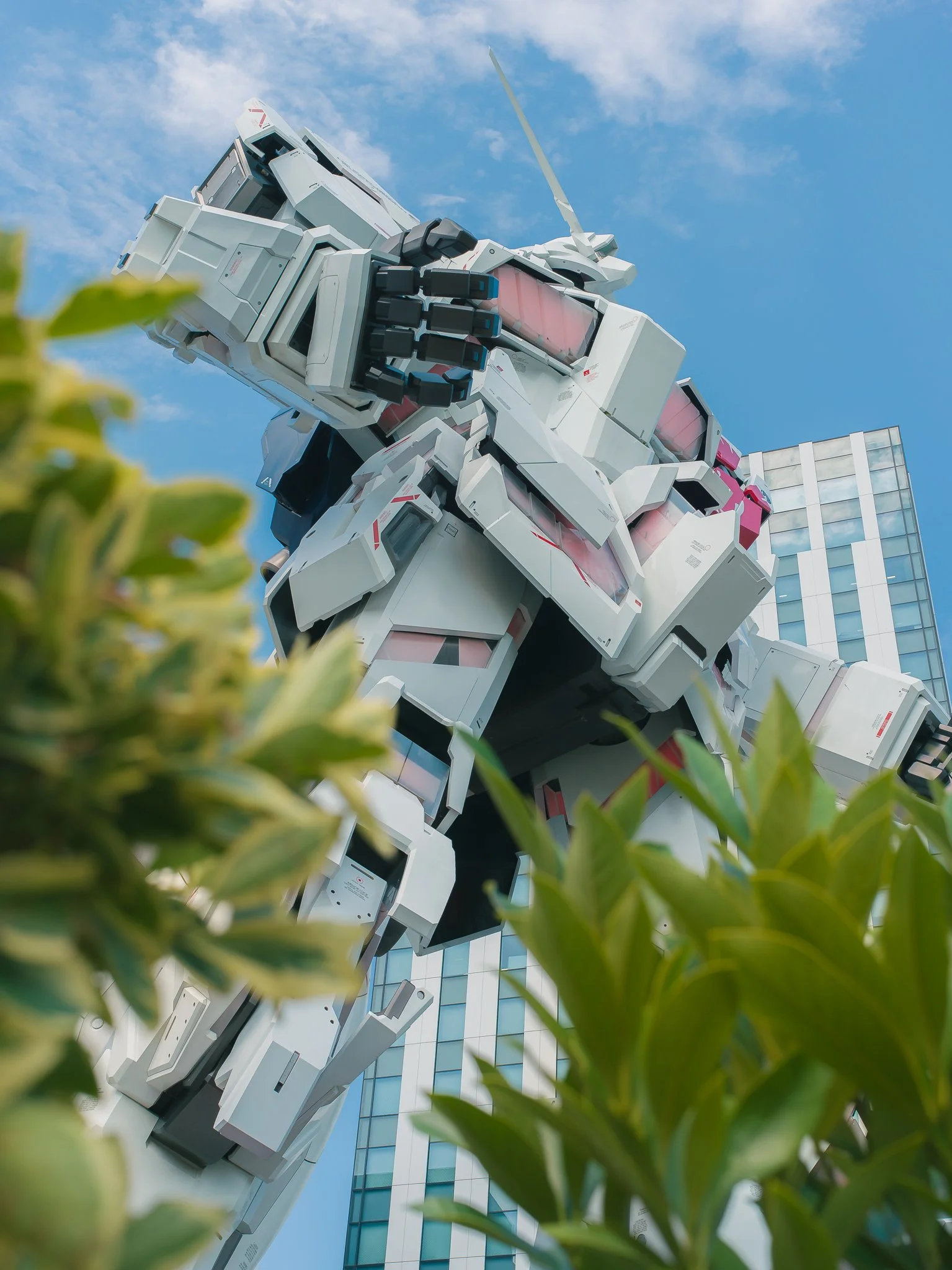 A large, white humanoid robot sculpture with black and pink accents standing outdoors with a modern glass building and a blue sky with scattered clouds in the background, surrounded partially by green leaves in the foreground.