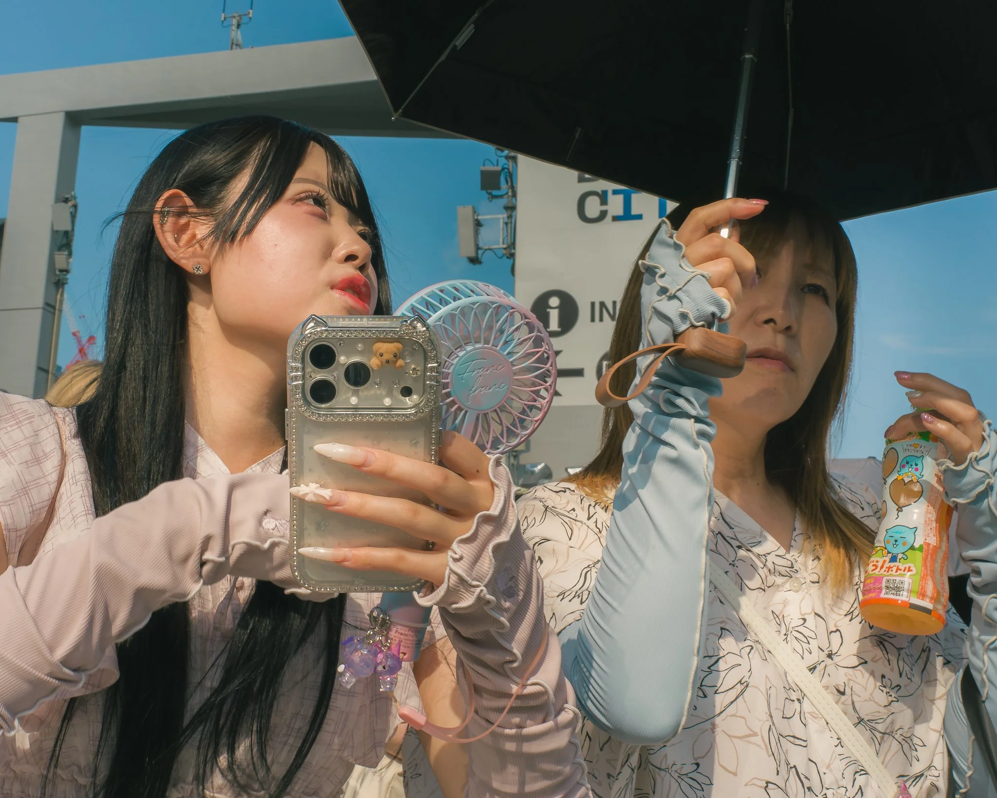 Two women standing outdoors under a black umbrella, one holding a smartphone with a bear decoration on the case, and the other holding a snack. Both are wearing patterned clothing and have long hair, with a blue sky and signs visible in the backgroun