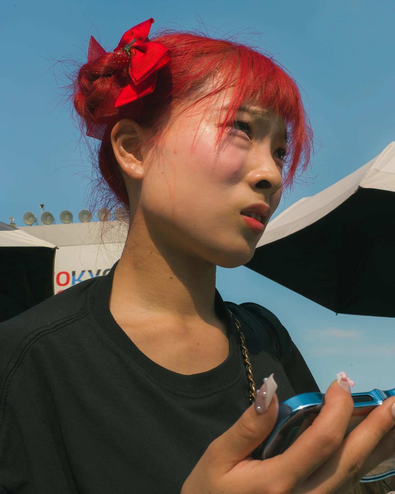 A woman with red hair adorned with a red bow, holding a smartphone, outdoors with umbrellas and a clear blue sky in the background.