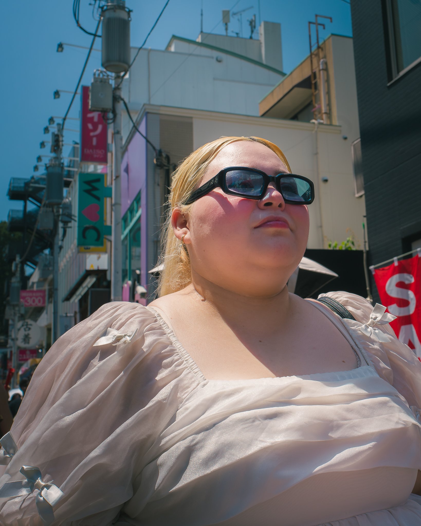 A woman with blonde hair wearing black sunglasses and a beige top, standing outdoors in an urban area with buildings and power lines in the background.