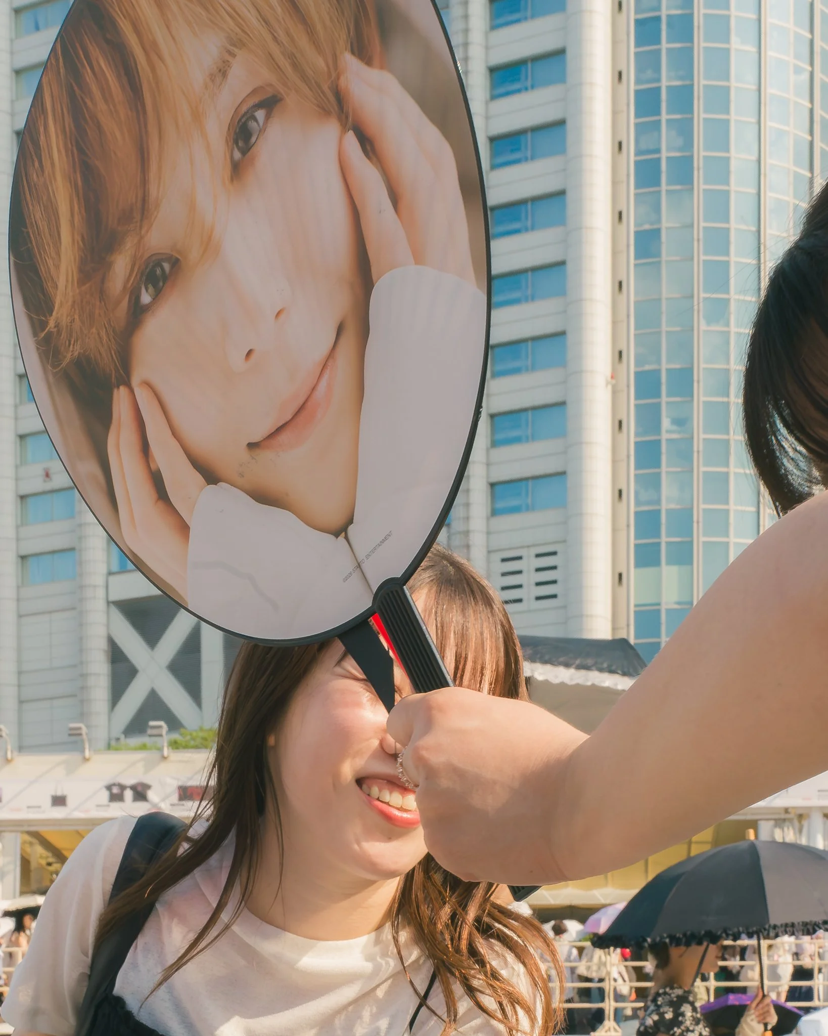 A woman with long brown hair smiling while holding a fan with a large image of a girl's face on it, in an outdoor urban setting with buildings and people in the background.