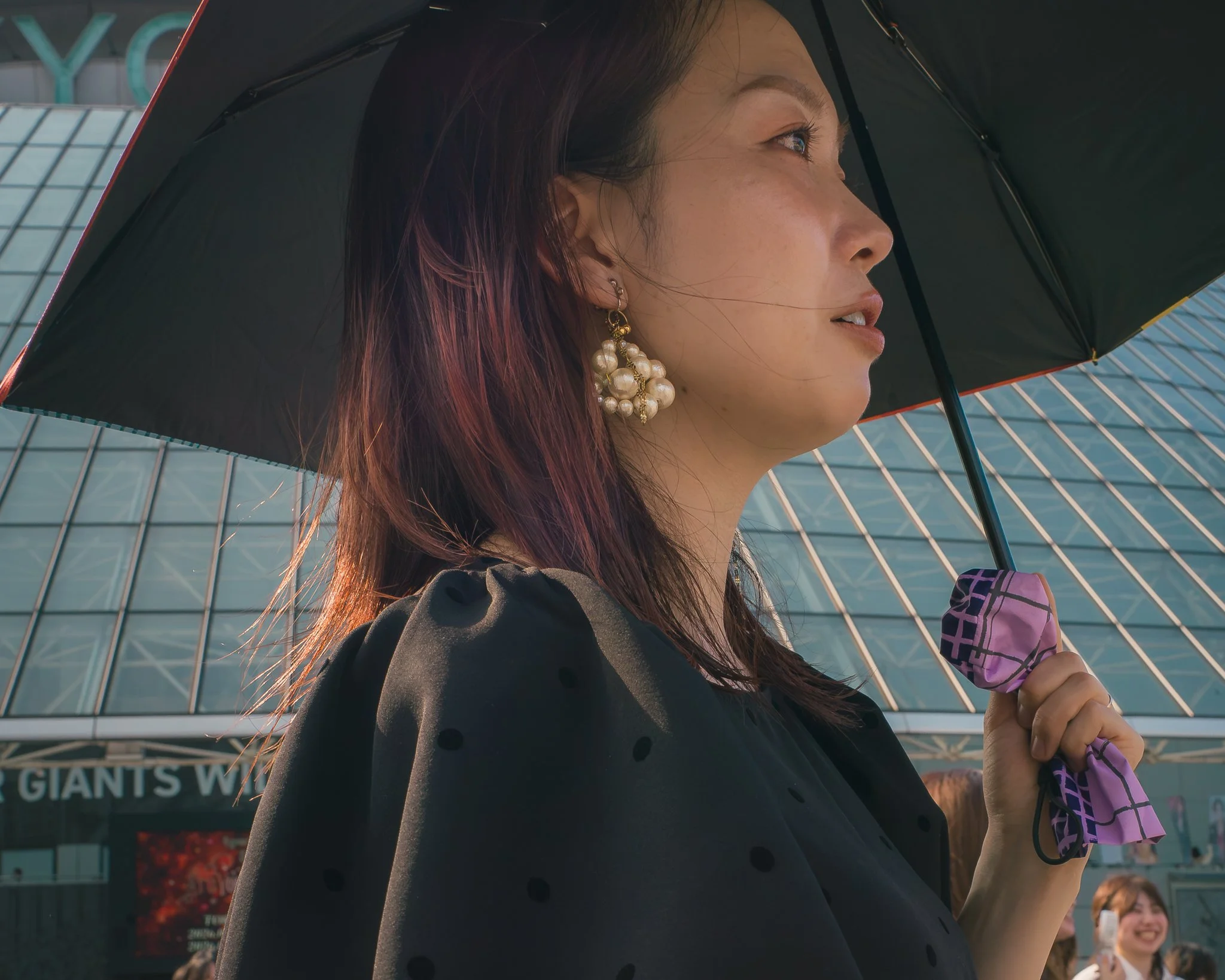 Close-up side profile of a woman with dark hair, wearing large pearl earrings, carrying a black umbrella, in an urban setting with a modern glass building in the background.