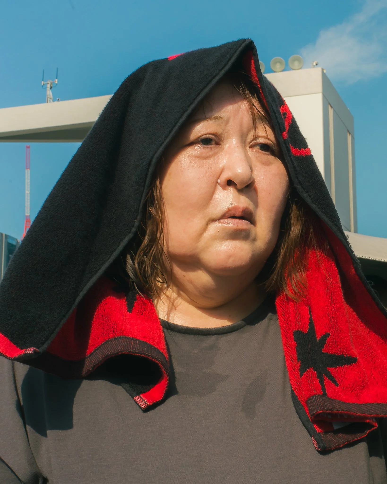 Close-up of a woman with wet hair and a black and red scarf, standing outdoors with a blue sky and communication towers in the background.