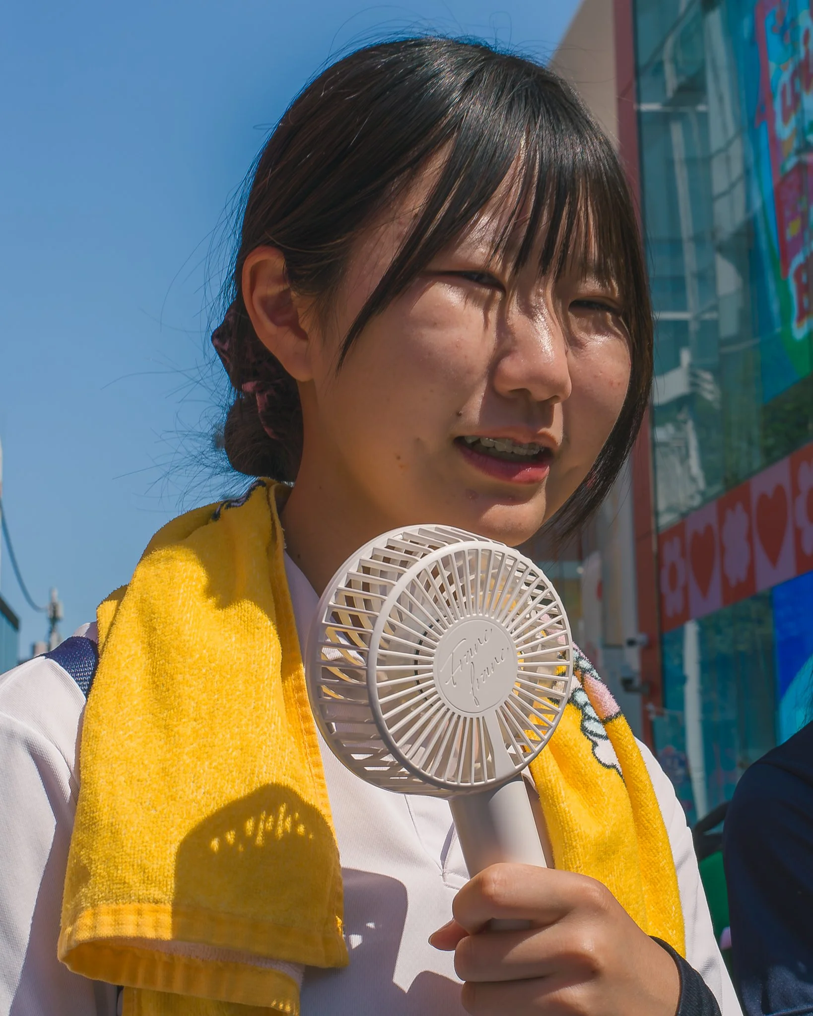 Close-up of a woman holding a white handheld fan, wearing a white shirt and a yellow towel draped over her shoulder, with a background of a billboard and a clear blue sky.