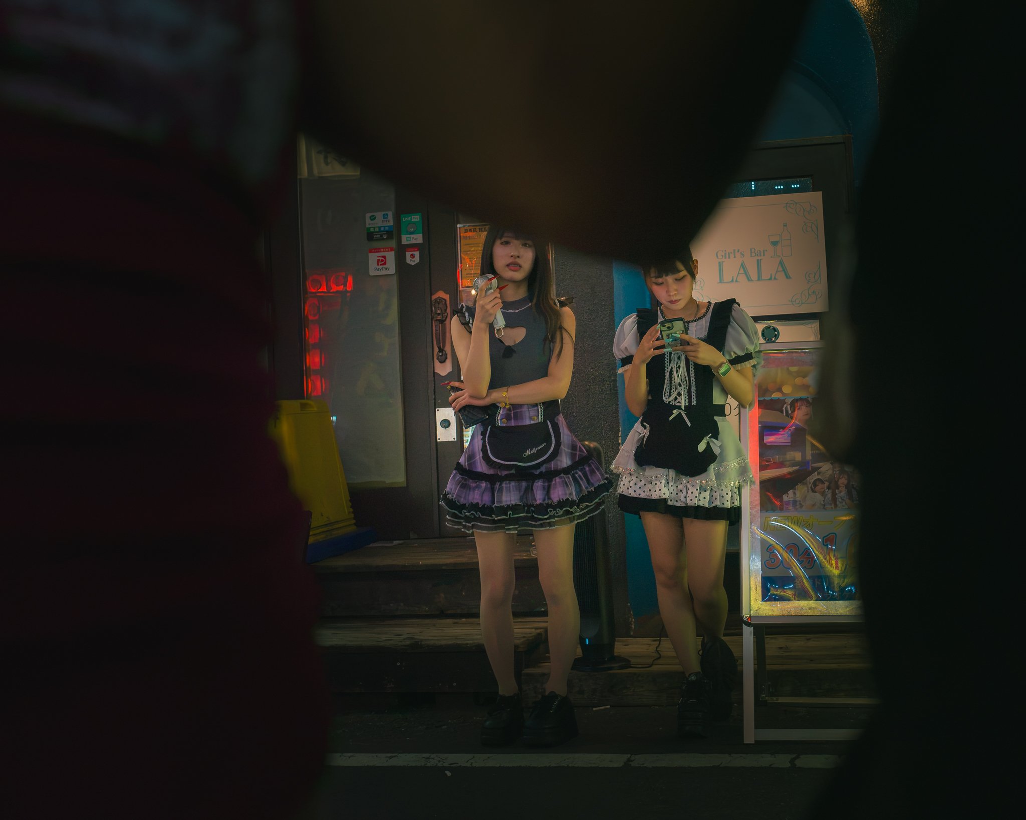 Two young women standing in front of a sign that reads 'Girl's Bar LALA'. One woman is looking at her phone, and the other is holding a microphone.