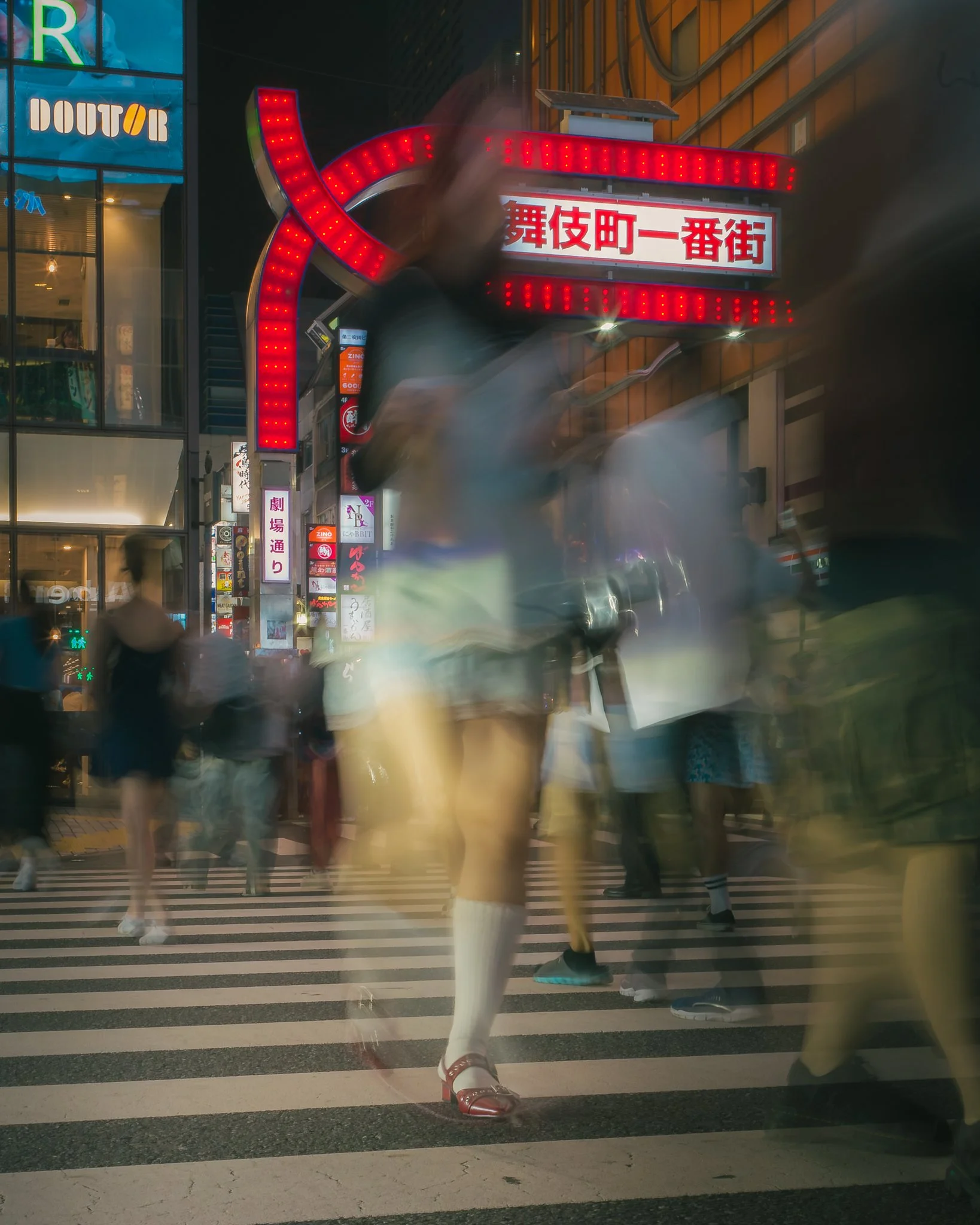 Nighttime street scene with illuminated signs and blurred pedestrians crossing the street. Visible Japanese characters on signs and advertisements for various businesses.
