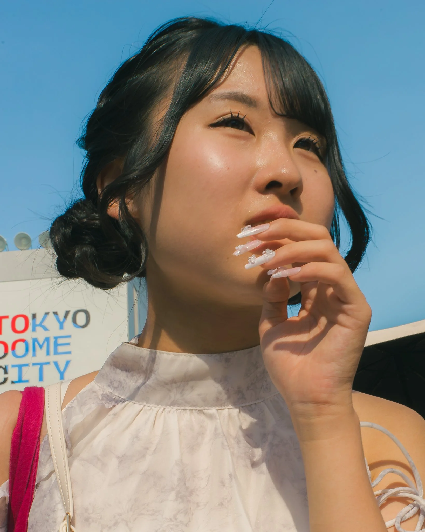 Close-up of a woman with black hair styled in a bun, with some loose bangs, looking into the distance against a blue sky, wearing a white top with a pink strap on her shoulder, and a white bag with a gold zipper.