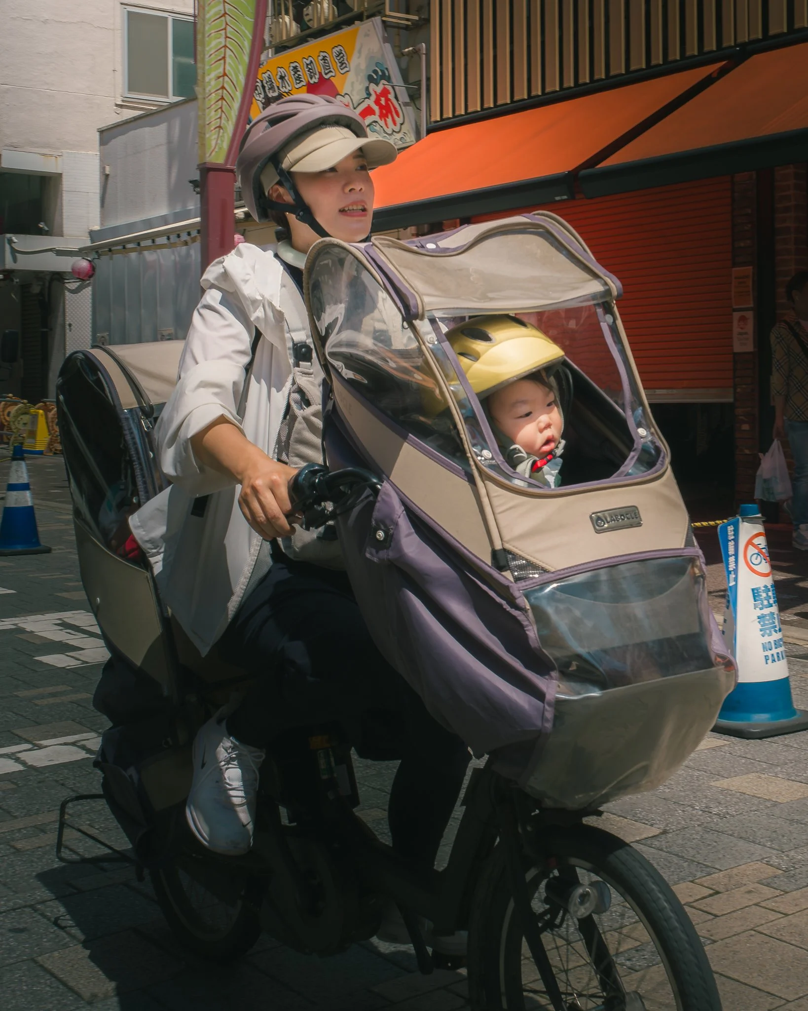 A woman wearing a beige helmet and white jacket riding a tricycle with a child in a beige stroller. The stroller has a clear plastic cover and the woman is smiling, riding on a city street with orange and yellow storefronts.