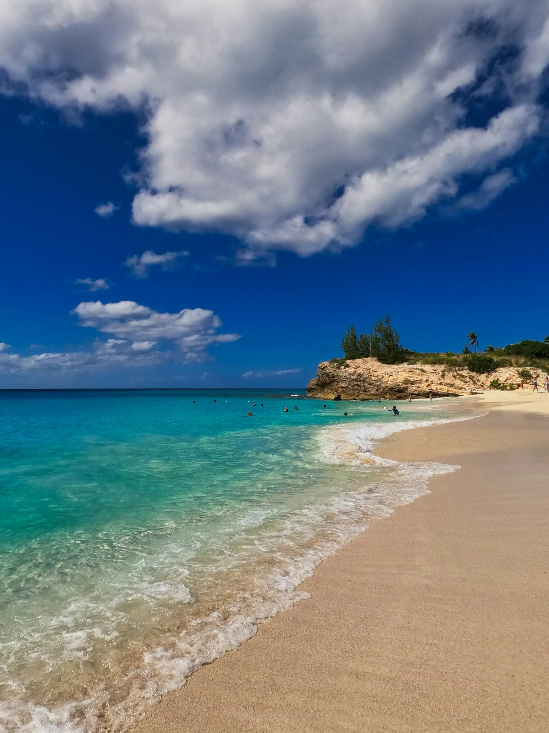 Beach with turquoise water, sandy shoreline, rocky cliff, and cloudy sky, people swimming in the ocean.