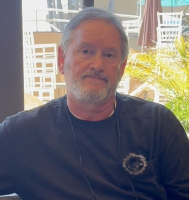 Man with gray hair and beard wearing a black shirt sitting indoors with sunlight, plants, and outdoor furniture visible in the background.
