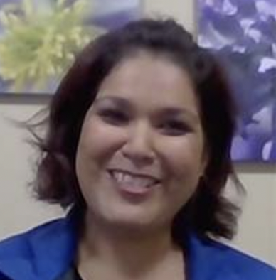 A woman smiling, with shoulder-length dark hair, wearing a blue jacket, in front of a background with plants and purple leaves.