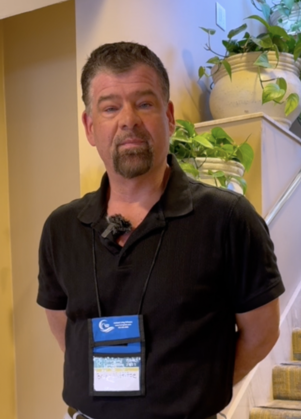 Man with short brown hair, beard, and mustache, wearing a black polo shirt and a conference badge around his neck, standing indoors near a staircase with green potted plants on the side.