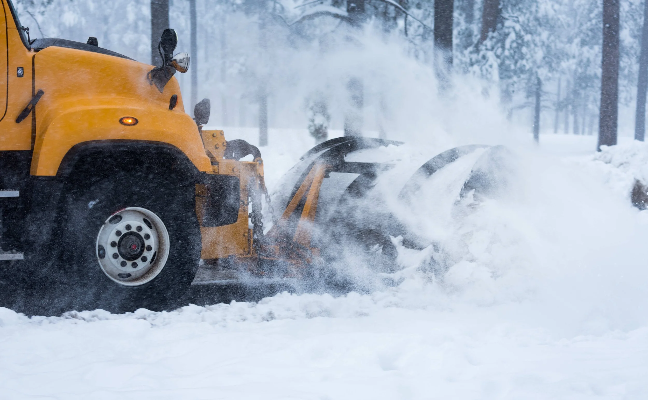 A yellow snow plow clearing snow from a road in a snowy forest.