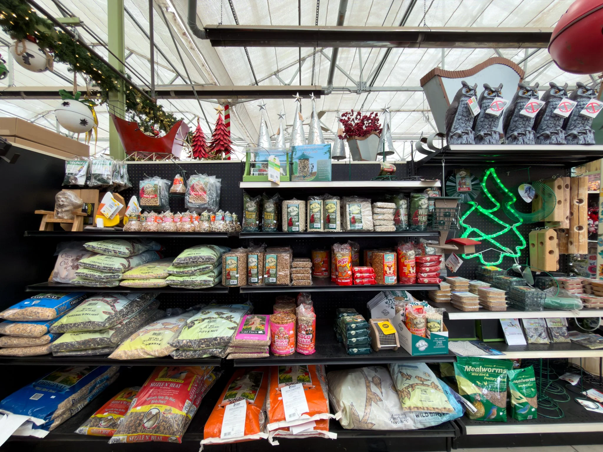 Shelf display of bird seed and feed products in a store, decorated with Christmas ornaments, including a neon green Christmas tree sign and animal figurines.