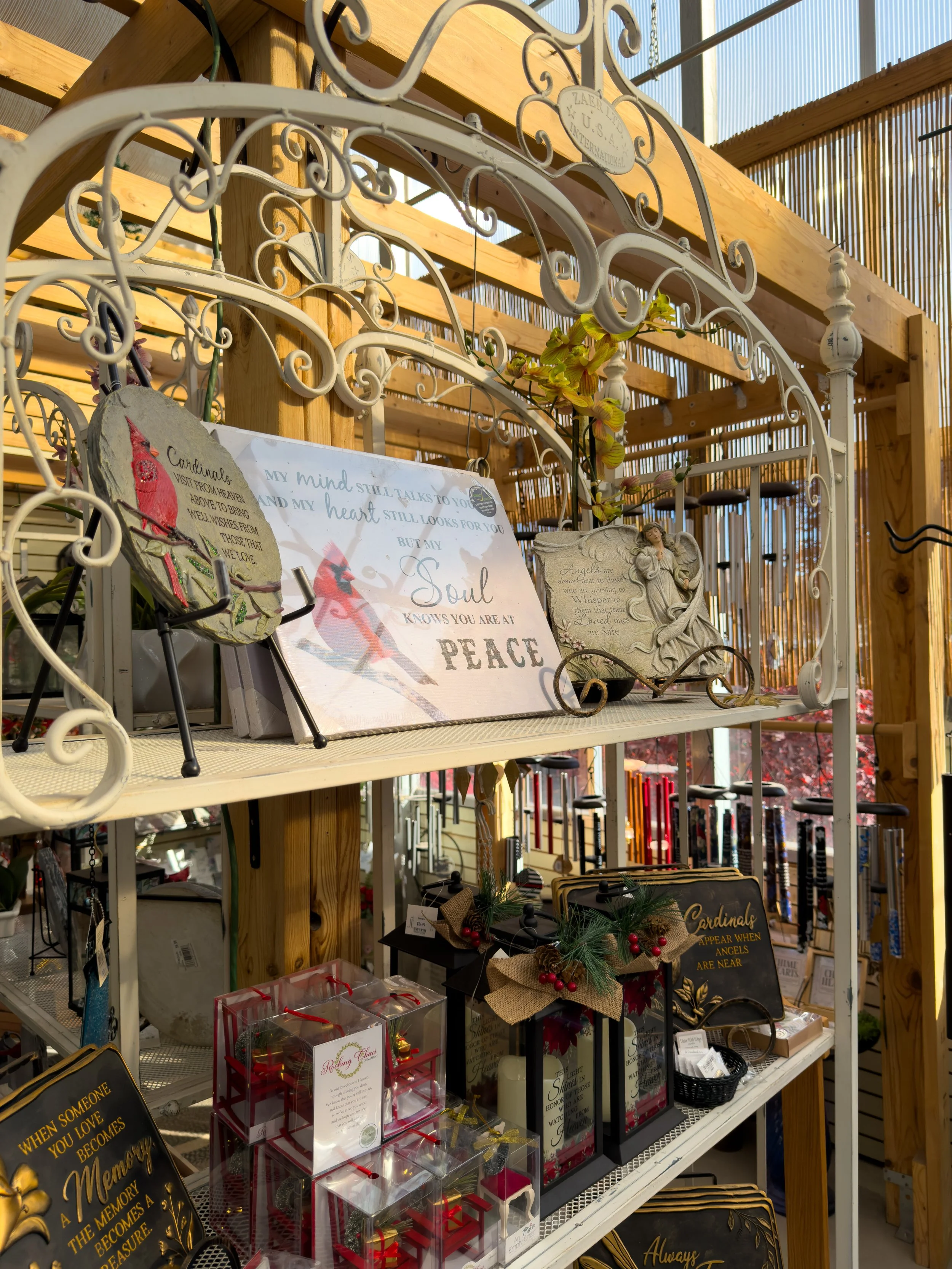 Decorative garden display with angel and nature-themed signs, ornaments, and gift items on metal and wooden shelves, with a wooden fence and outdoor sky in the background.