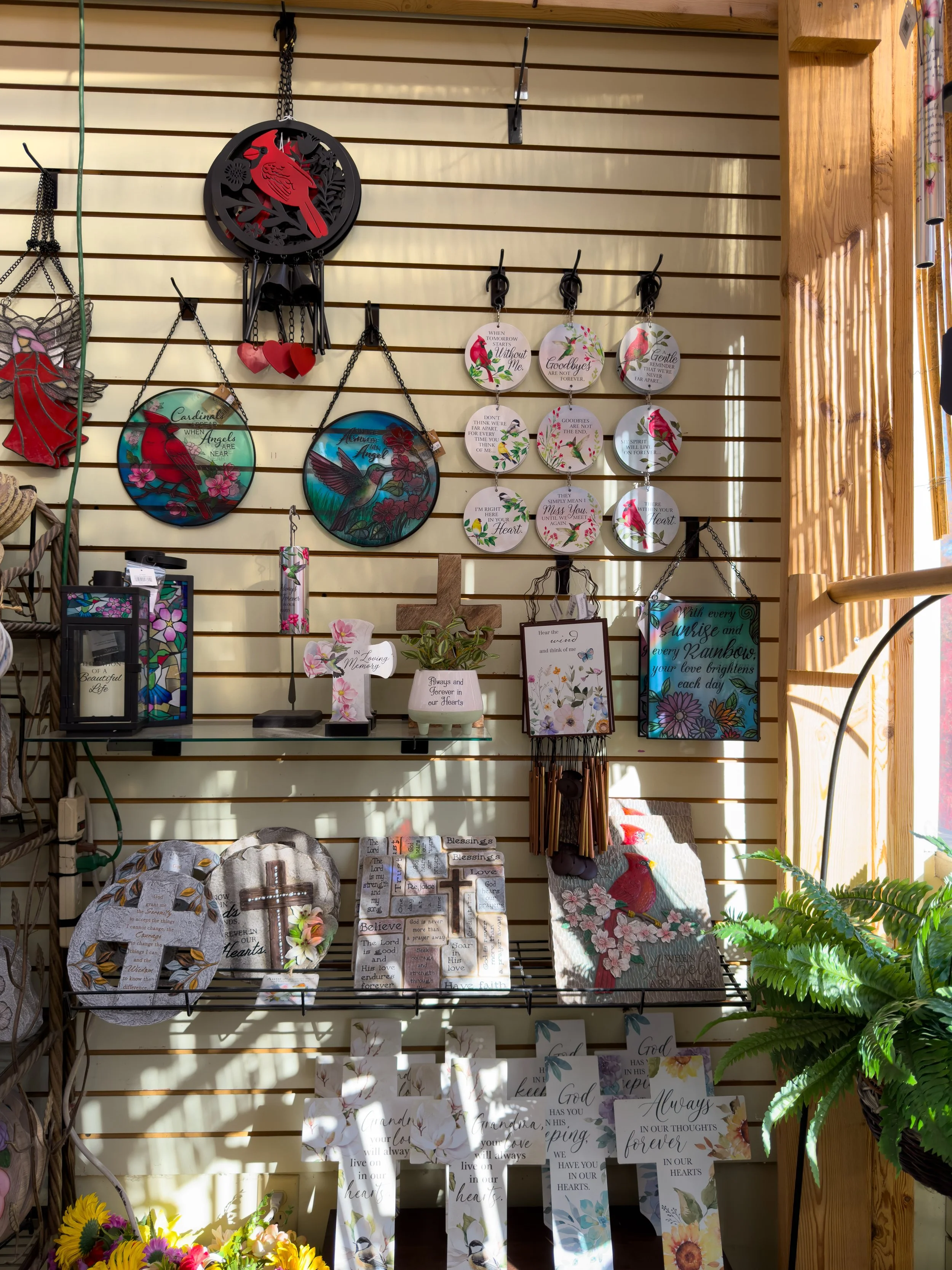 Display of decorative items including circular stained glass artworks, plaques with sentimental quotes, crosses, a small potted plant, a red cardinal figurine, a floral picture, and sunflower arrangements on shelves and hooks.
