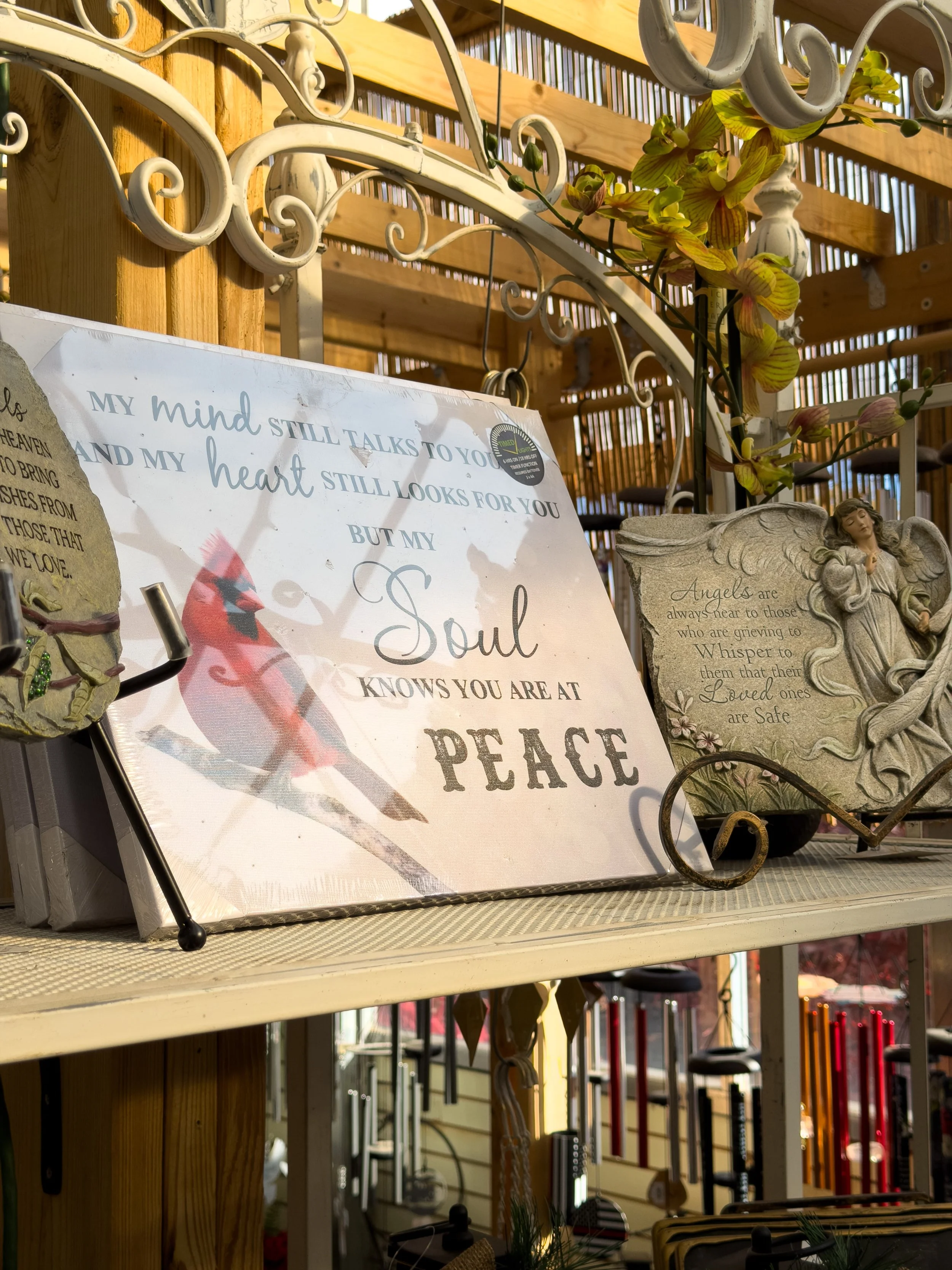 Decorative outdoor display with faith-inspired items including a sign with the message about the soul and peace, along with angel figurines and artificial flowers, set against a wooden fence and lattice background.
