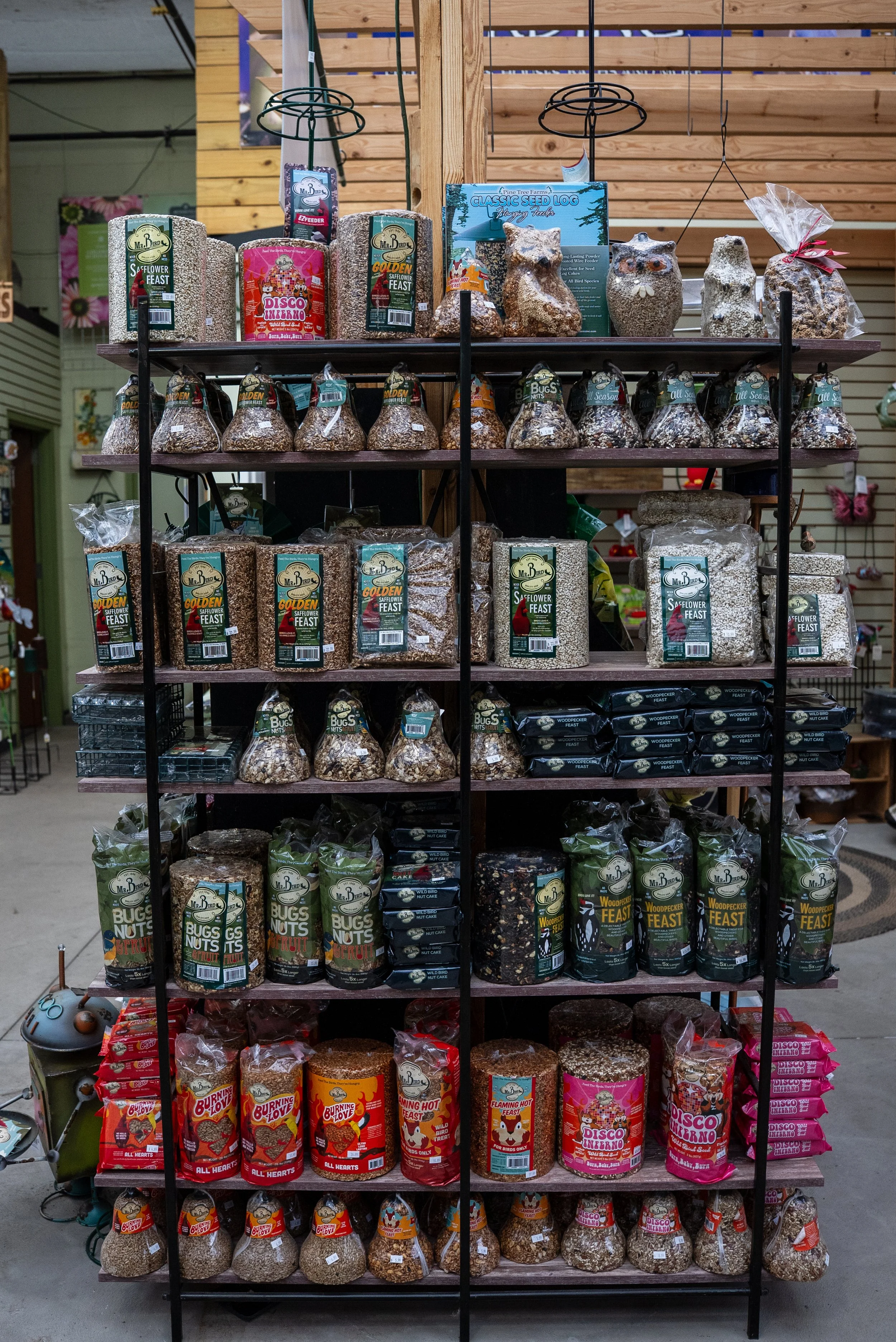 Display of various birdseed and bird food in a store, arranged on a multi-level shelf with different packaging and product types.