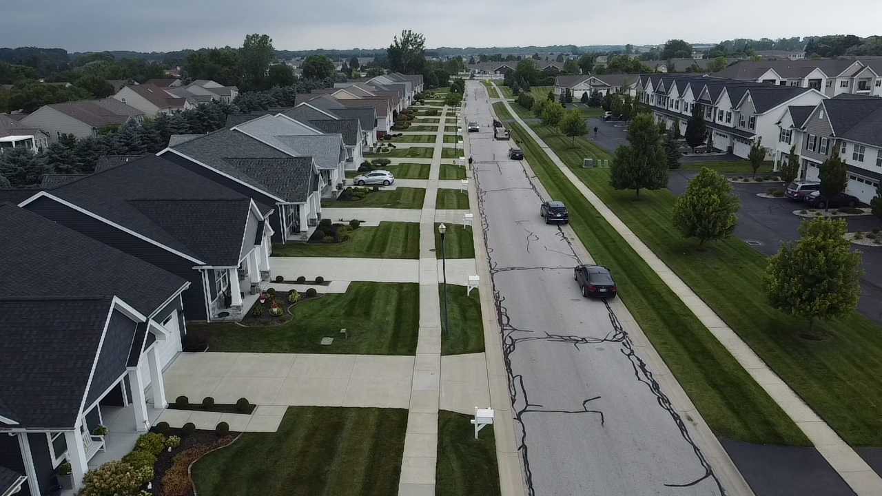 A residential neighborhood with single-family homes, manicured lawns, sidewalks, and parked cars along a crack-filled street under a cloudy sky.