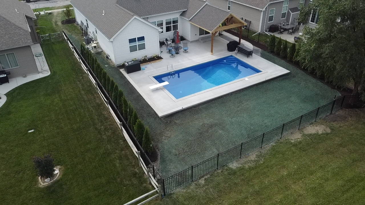 A man wearing a black hoodie with the word 'RELIABLE' on the back is spraying green liquid on a dirt area in a backyard, with a small beige shed and larger white house with gray roof in the background on a sunny day with clear blue sky.