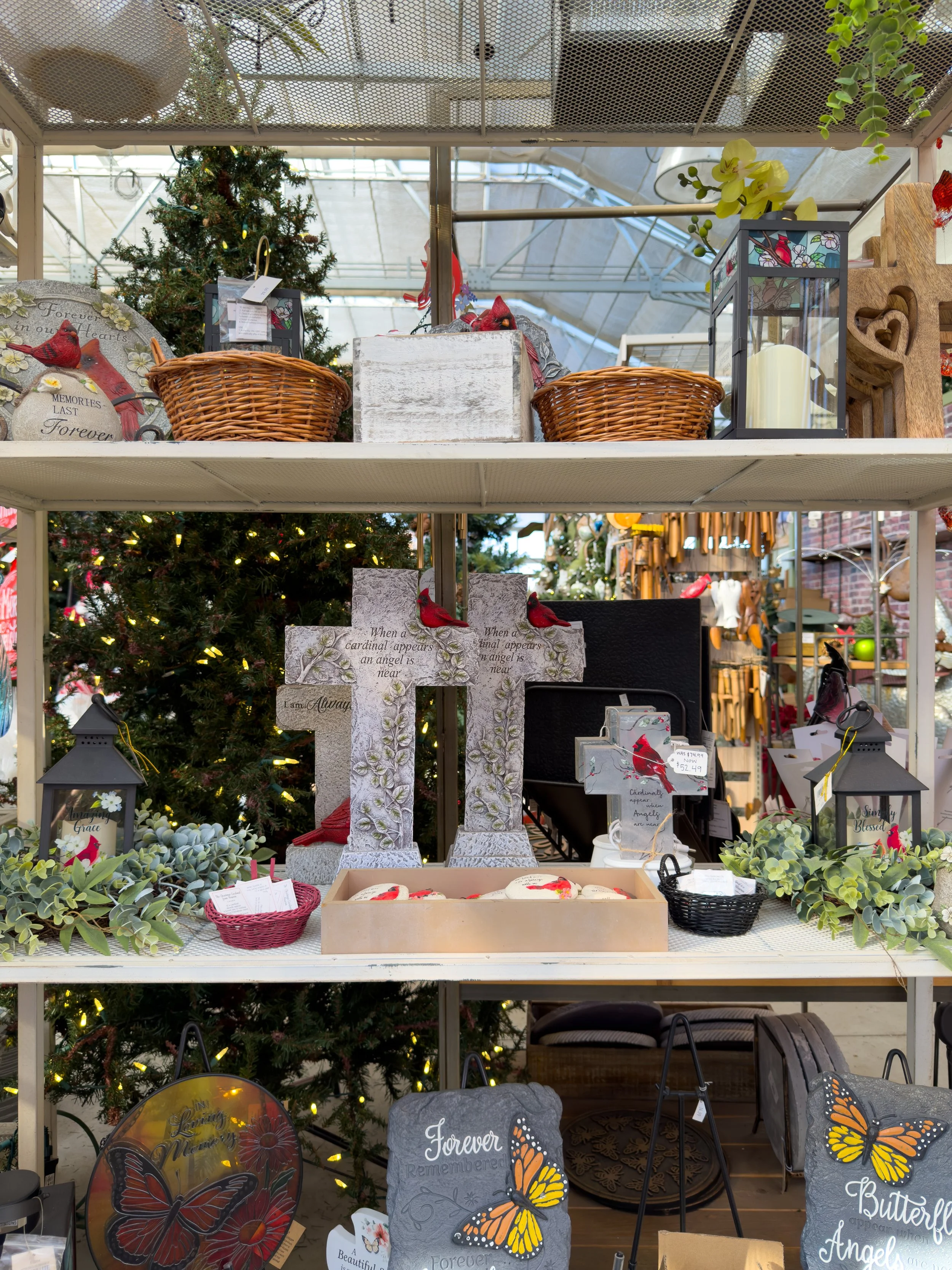 Decorative items on a store shelf, including a small Christmas tree, woven baskets, candles, crosses with red cardinal birds, lanterns, and butterfly-themed pillows with greenery.