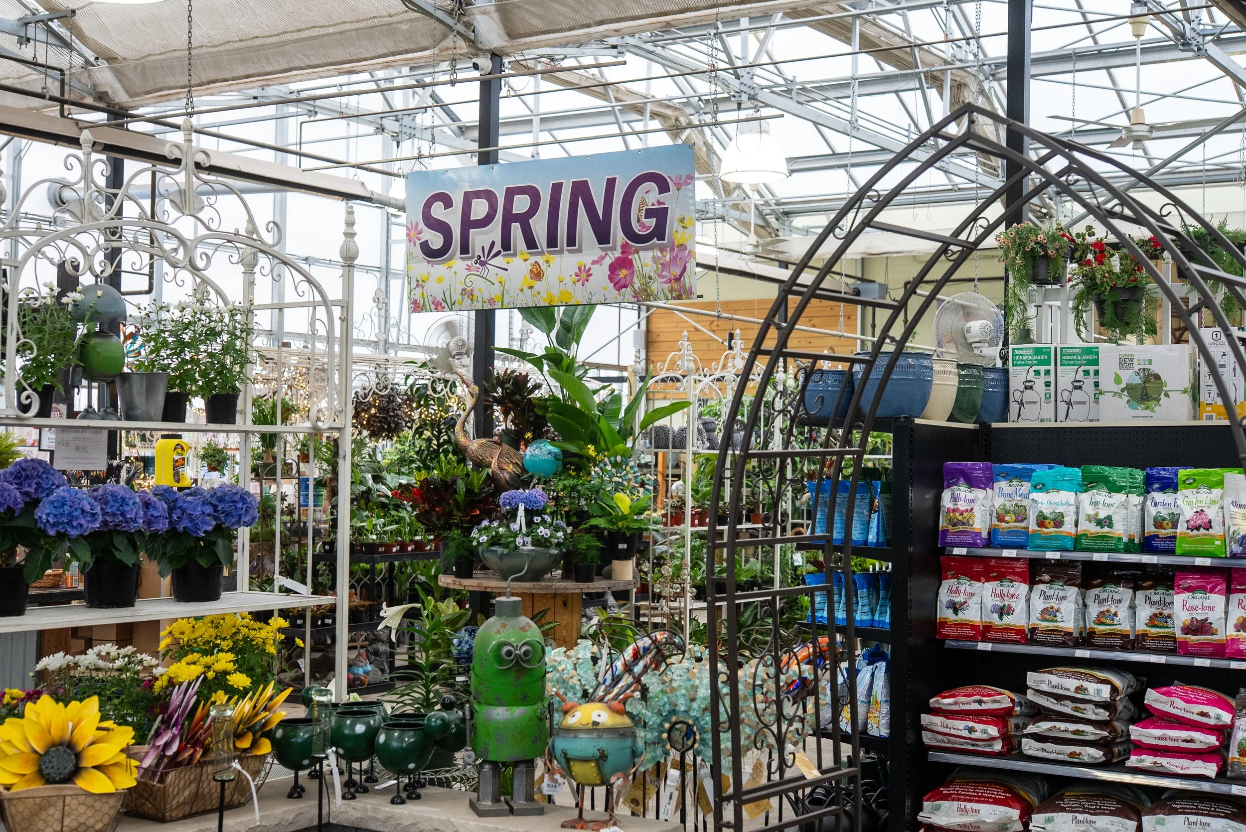 A display of spring-themed garden and plant items inside a greenhouse, including potted flowers, garden decor, and gardening supplies, under a sign that reads 'Spring'.