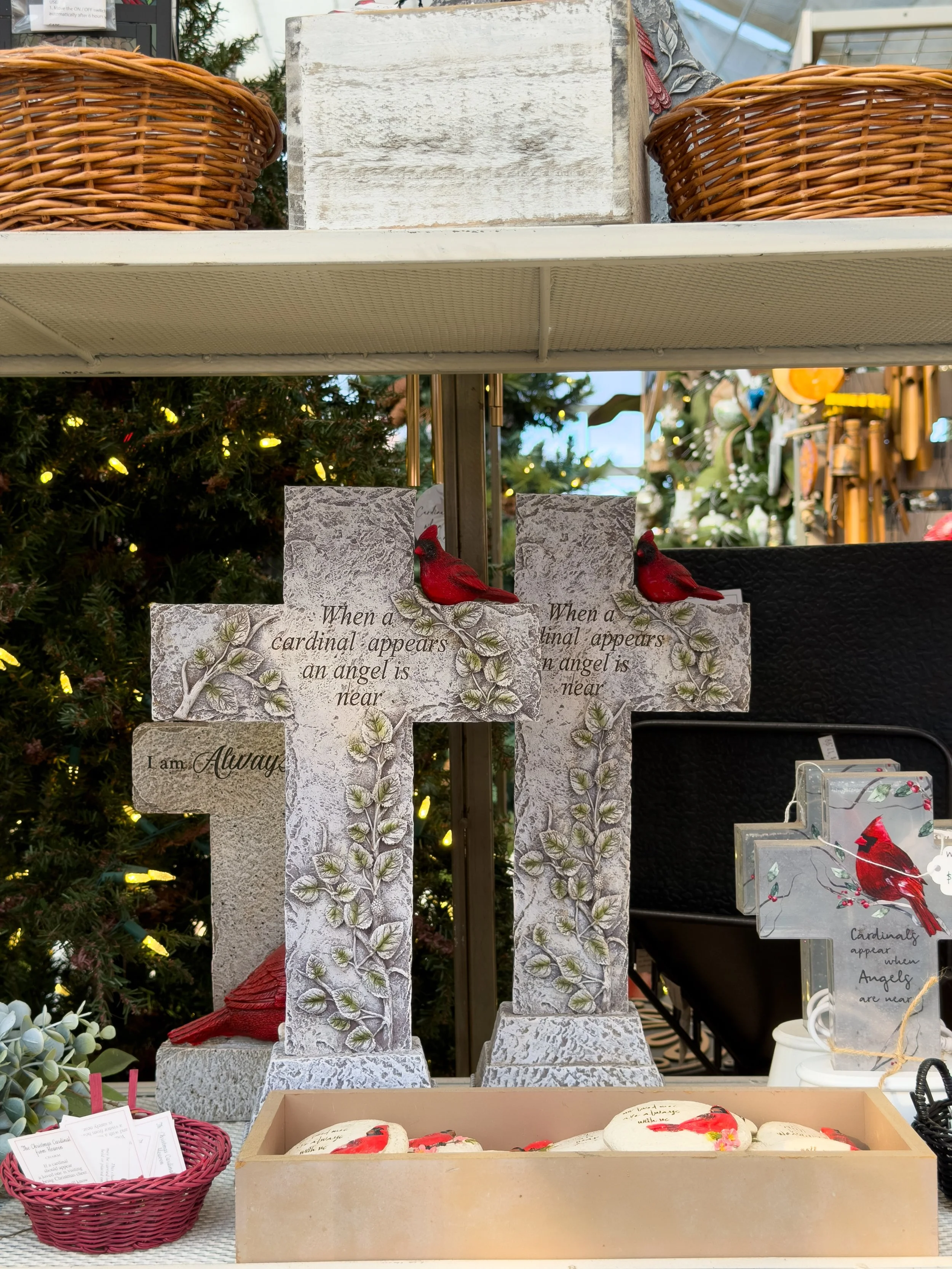 Decorative stone crosses with red cardinal bird ornaments and inspirational quotes, Christmas tree in the background, pottery and baskets displayed on shelves.