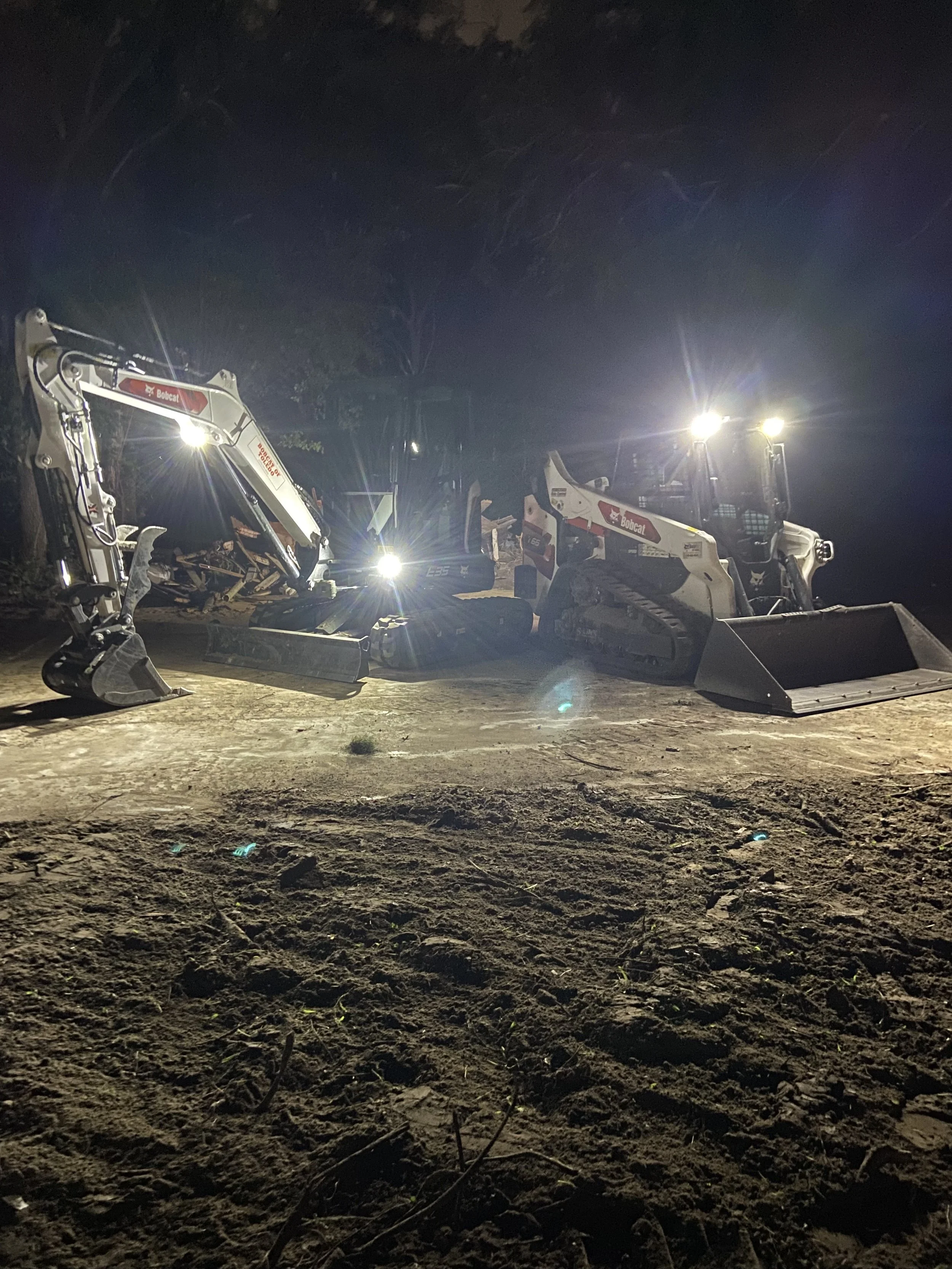 Nighttime scene with two small construction vehicles, specifically Bobcat excavators, on dirt ground with lights turned on illuminating the area, and trees in the background.