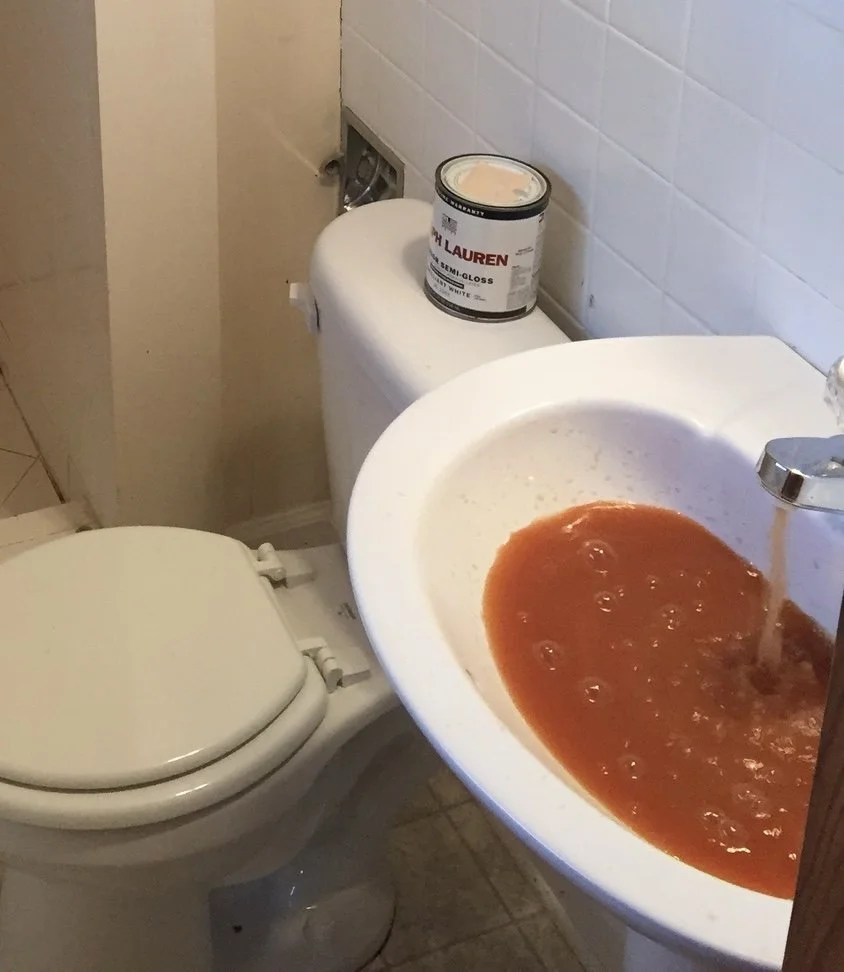 A small bathroom with a toilet and sink, with tomato sauce or juice in the sink and a can of semi-gloss beige paint on the toilet tank, near the wall.