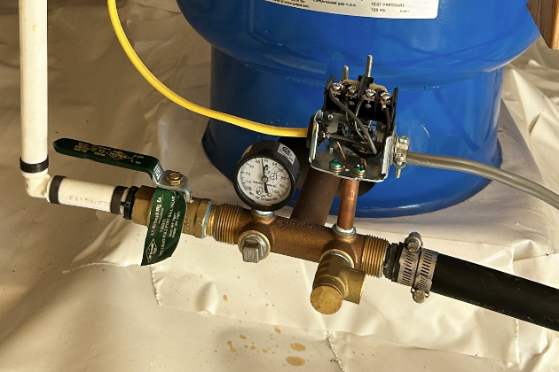 A plumbing setup with a pressure gauge, a valve, and various pipes connected to a large blue tank in the background.