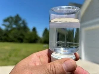 Hand holding a clear water sample outdoors before laboratory submission for bacteria testing.