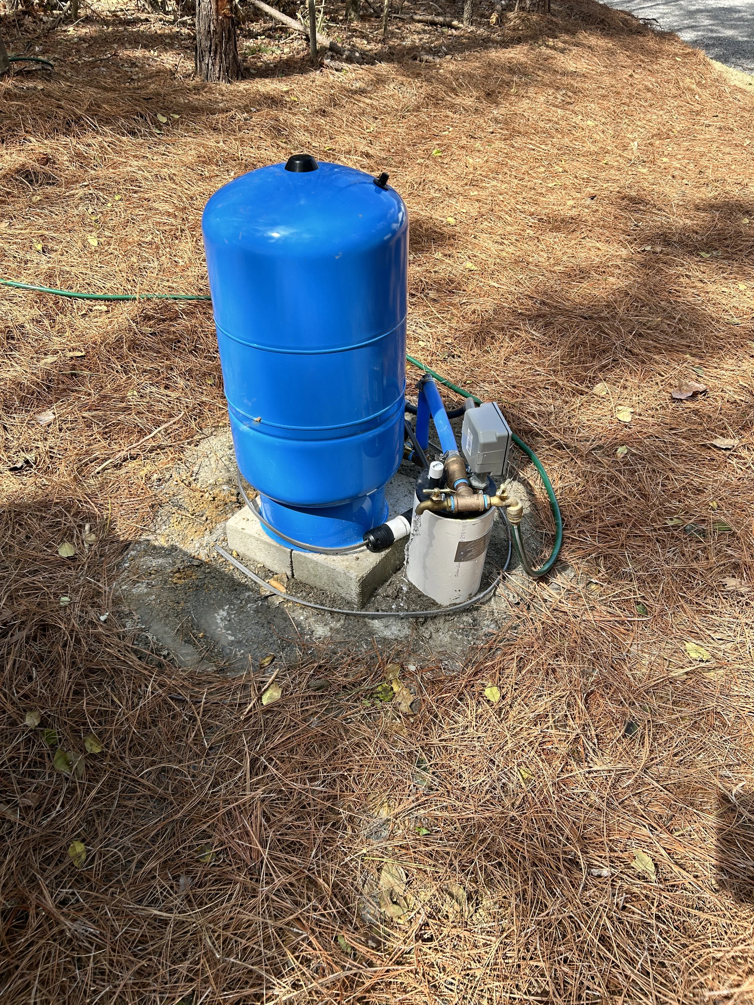 Blue outdoor water pump with pipes, situated on a concrete block in a wooded area with pine needles and trees.
