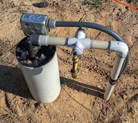 A water filtration system with a white cylindrical filter, connected to gray and black pipes, installed outdoors on dirt ground.