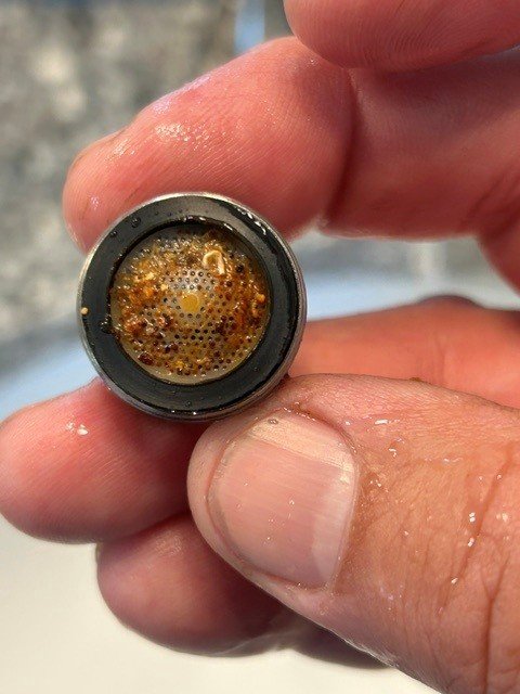 Hand holding a faucet aerator with orange-brown deposits on its mesh screen.