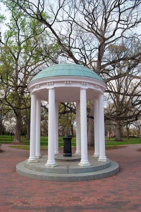 A classical white pavilion with a green domed roof, supported by eight tall columns, situated on a circular brick platform in a park with trees in the background.