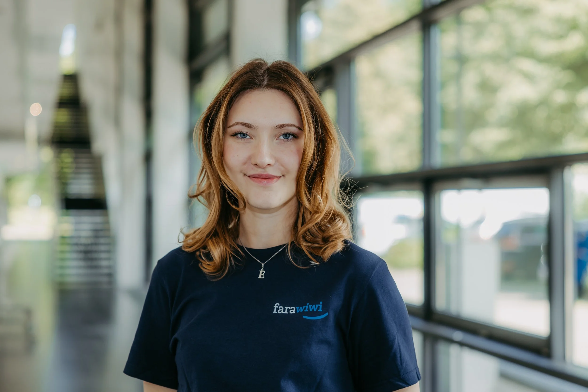 Junge Frau mit roten Haaren, blauer T-Shirt und Herzschmuck, lächelnd, im Hintergrund eines Gebäudes mit großen Fenstern und grüner Natur draußen.