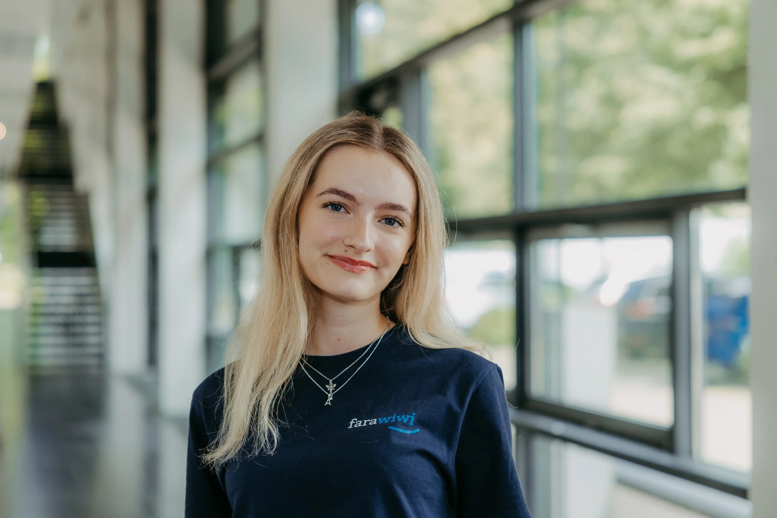 Junge Frau mit blondem Haar, trägt ein schwarzes T-Shirt mit dem Logo 'fara wiwi', steht in einem hellen, modernen Gebäude mit großen Fenstern und grüner Umgebung im Hintergrund.