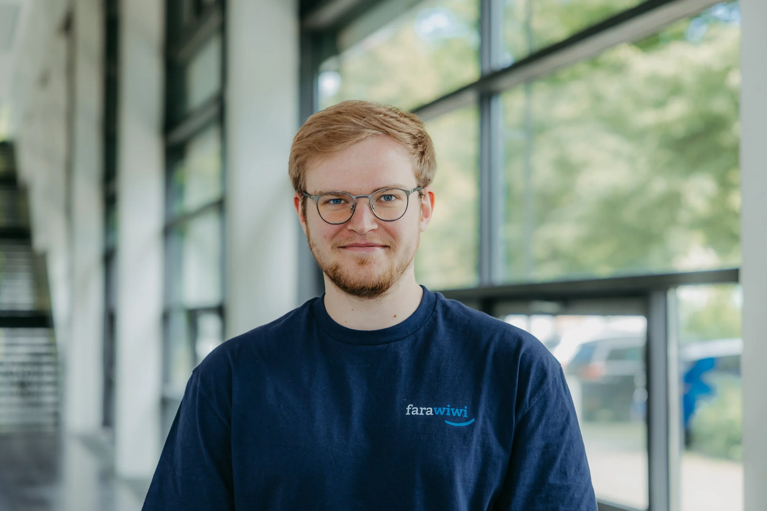 Junger Mann mit Brille trägt ein dunkelblaues T-Shirt mit Logo, steht in einem Raum mit großen Fenstern und grüner Natur im Hintergrund.