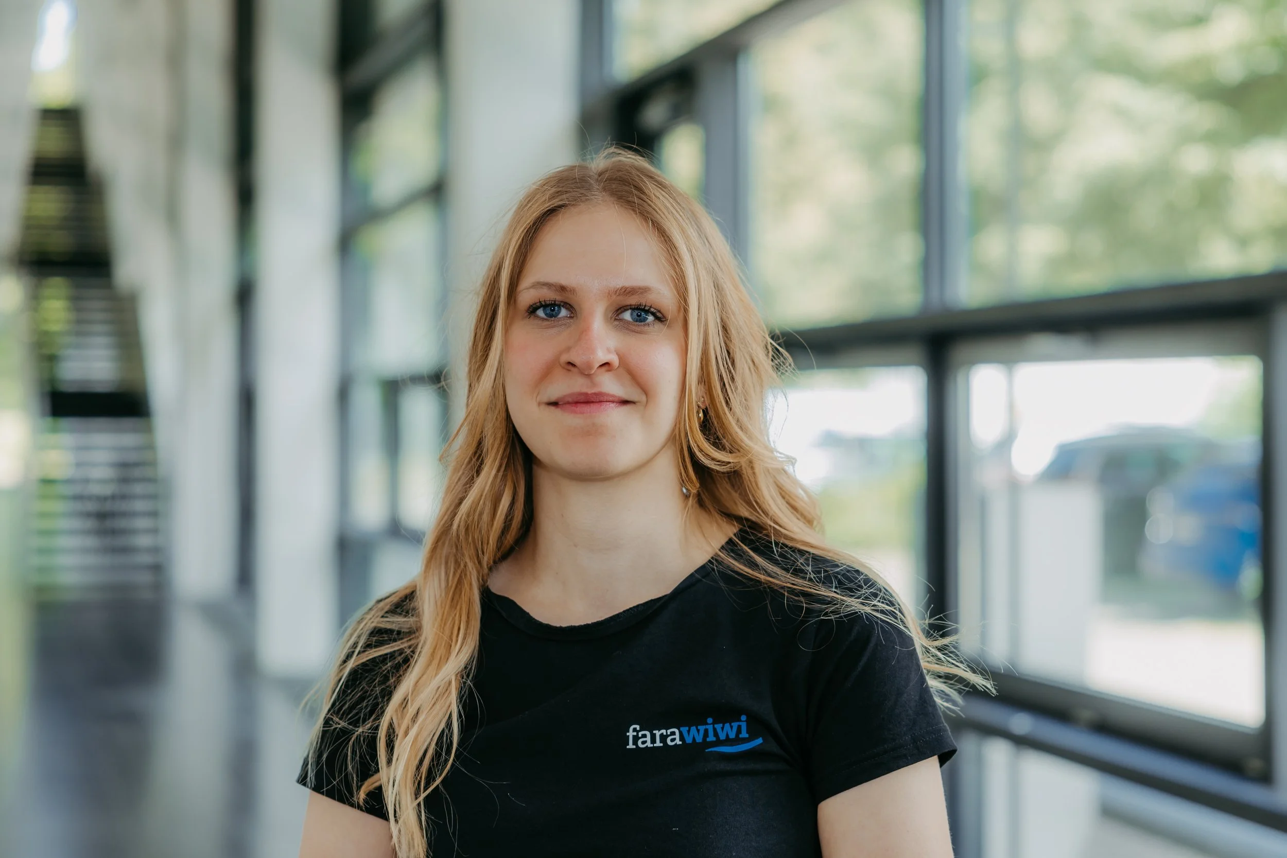 Junge Frau mit langen blonden Haaren und blauen Augen in einem schwarzen T-Shirt mit dem Logo 'fara wiwi', die in einem hell erleuchteten Raum mit großen Fenstern steht.